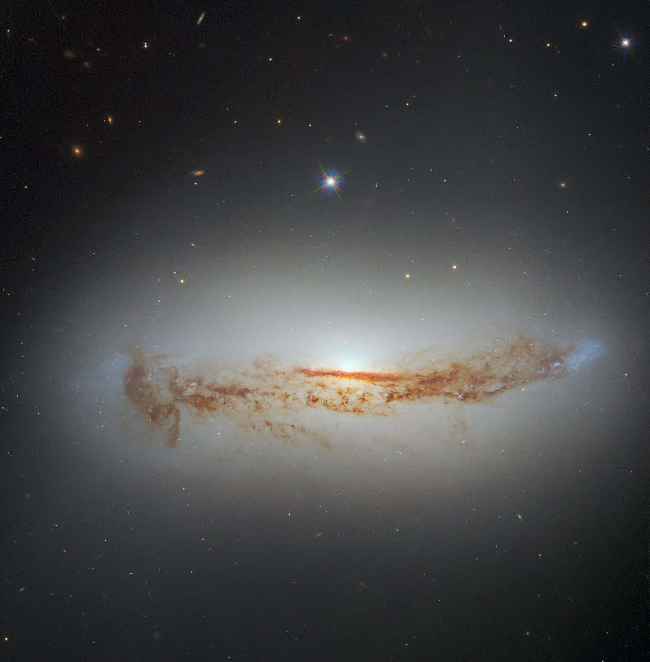 Tendrils of dark dust can be seen threading across the heart of the spiral galaxy NGC 7172 in this image from the NASA/ESA Hubble Space Telescope. The galaxy lies approximately 110 million light-years from Earth in the constellation Piscis Austrinus. The lane of dust threading its way across NGC 7172 — which is viewed side-on in this image — is obscuring the luminous heart of the galaxy, making NGC 7172 appear to be nothing more than a normal edge-on spiral galaxy.   When astronomers inspected NGC 7172 across the electromagnetic spectrum they quickly discovered that there was more to it than meets the eye: NGC 7172 is a Seyfert galaxy — a type of galaxy with an intensely luminous active galactic nucleus powered by matter accreting onto a supermassive black hole.   This image combines data from two sets of Hubble observations, both of which were proposed to study nearby active galactic nuclei. The image also combines data from two instruments — Hubble’s Advanced Camera for Surveys (ACS) and Wide Field Camera 3 (WFCS).  Credit: ESA/Hubble & NASA, D. J. Rosario, A. Barth  Acknowledgement: L. Shatz