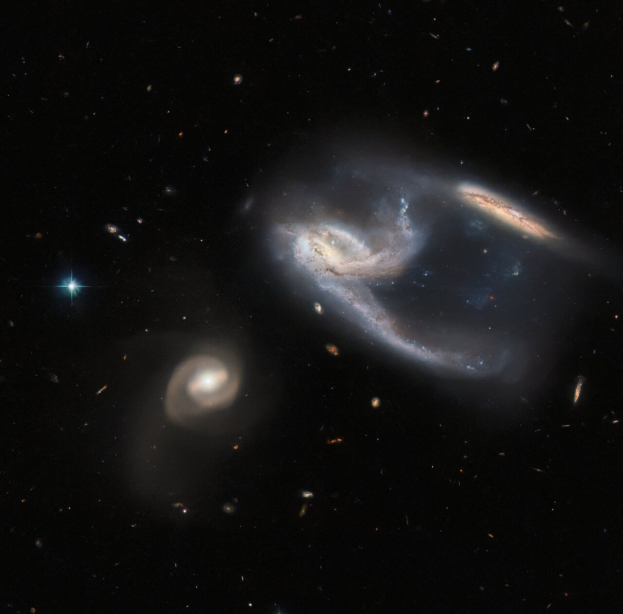 The subject of this image is a group of three galaxies, collectively known as NGC 7764A. They were imaged by the NASA/ESA Hubble Space Telescope, using both its Advanced Camera for Surveys (ACS) and Wide Field Camera 3 (WFC3). The two galaxies in the upper right of the image appear to be interacting with one another — indeed, the long trails of stars and gas extending from them both give the impression that they have both just been struck at great speed, thrown into disarray by the bowling-ball-shaped galaxy to the lower left of the image. In reality, however, interactions between galaxies happen over very long time periods, and galaxies rarely collide head-on with one another. It is also unclear whether the galaxy to the lower left is actually interacting with the other two, although they are so relatively close in space that it seems possible that they are. By happy coincidence, the collective interaction between these galaxies have caused the two on the upper right to form a shape, which from our Solar System's perspective, ressembles the starship known as the USS Enterprise from Star Trek!  NGC 7764A, which lies about 425 million light years from Earth in the constellation Phoenix, is a fascinating example of just how awkward astronomical nomenclature can be. The three galaxies are individually referred to as NGC 7764A1, NGC 7764A2 and NGC 7764A3, and just to be really difficult, an entirely separate galaxy, named NGC 7764, sits in the skies about a Moon’s distance (as seen from Earth) away. This rather haphazard naming makes more sense when we consider that many of the catalogues for keeping track of celestial bodies were compiled well over 100 years ago, long before modern technology made standardising scientific terminology much easier. As it is, many astronomical objects have several different names, or might have names that are so similar to other objects’ names that they cause confusion.  Credit: ESA/Hubble & NASA, J. Dalcanton, Dark Energy Survey, DOE, FNAL, DECam, CTIO, NOIRLab/NSF/AURA, ESO Acknowledgement: J. Schmidt