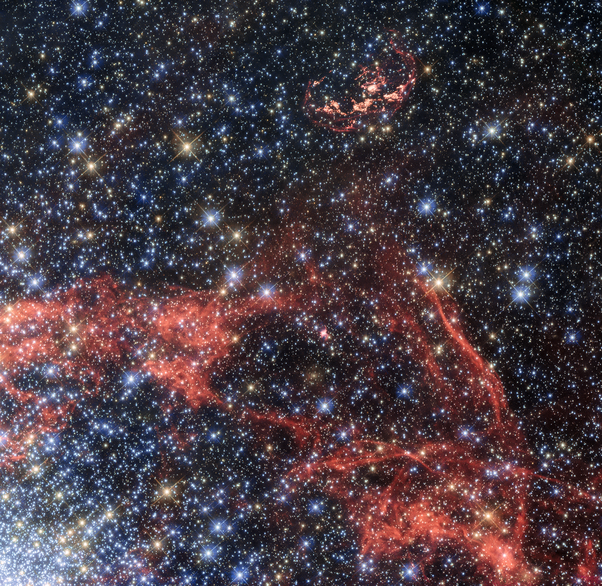 This image, taken with the NASA/ESA Hubble Space Telescope, shows the supernova remnant SNR 0509-68.7, also known as N103B (top of the image). N103B was a Type Ia supernova, located in the Large Magellanic Cloud — a neighbouring galaxy of the Milky Way. Owing to its relative proximity to Earth, astronomers observe the remnant to search for a potential stellar survivor of the explosion.  The orange-red filaments visible in the image show the shock fronts of the supernova explosion. These filaments allow astronomers to calculate the original centre of the explosion. The filaments also show that the explosion is no longer expanding as a sphere, but is elliptical in shape. Astronomers assume that part of material ejected by the explosion hit a denser cloud of interstellar material, which slowed its speed. The shell of expanding material being open to one side supports this idea.  The gas in the lower half of the image and the dense concentration of stars in the lower left are the outskirts of the star cluster NGC 1850, which has been observed by Hubble in the past [heic0108].  Credit: ESA/Hubble, NASA