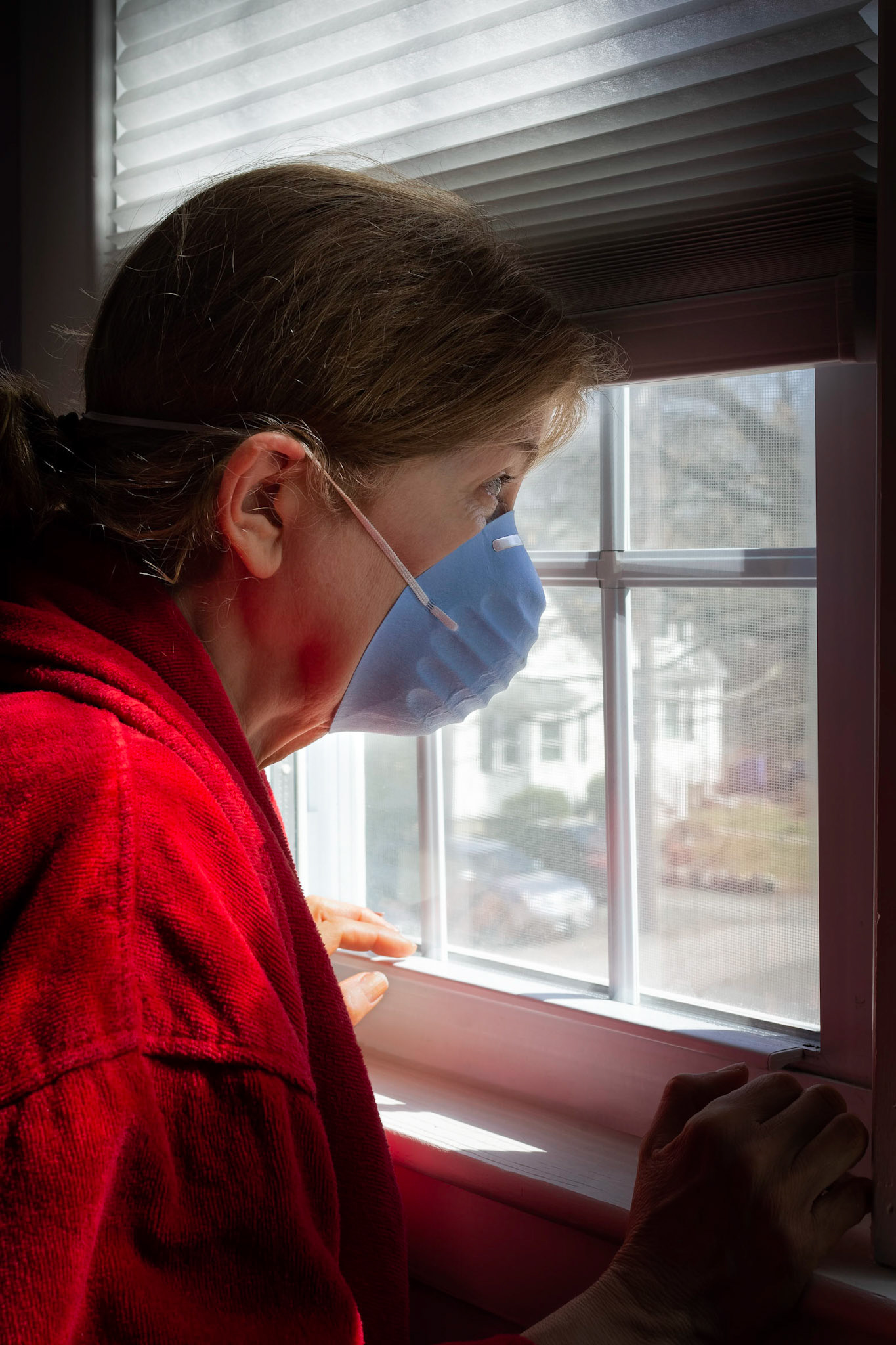 A woman looks longingly out a bright sunny window as if she wishes she could be outdoors while wearing a medical mask - COVID-19 (Coronavirus)