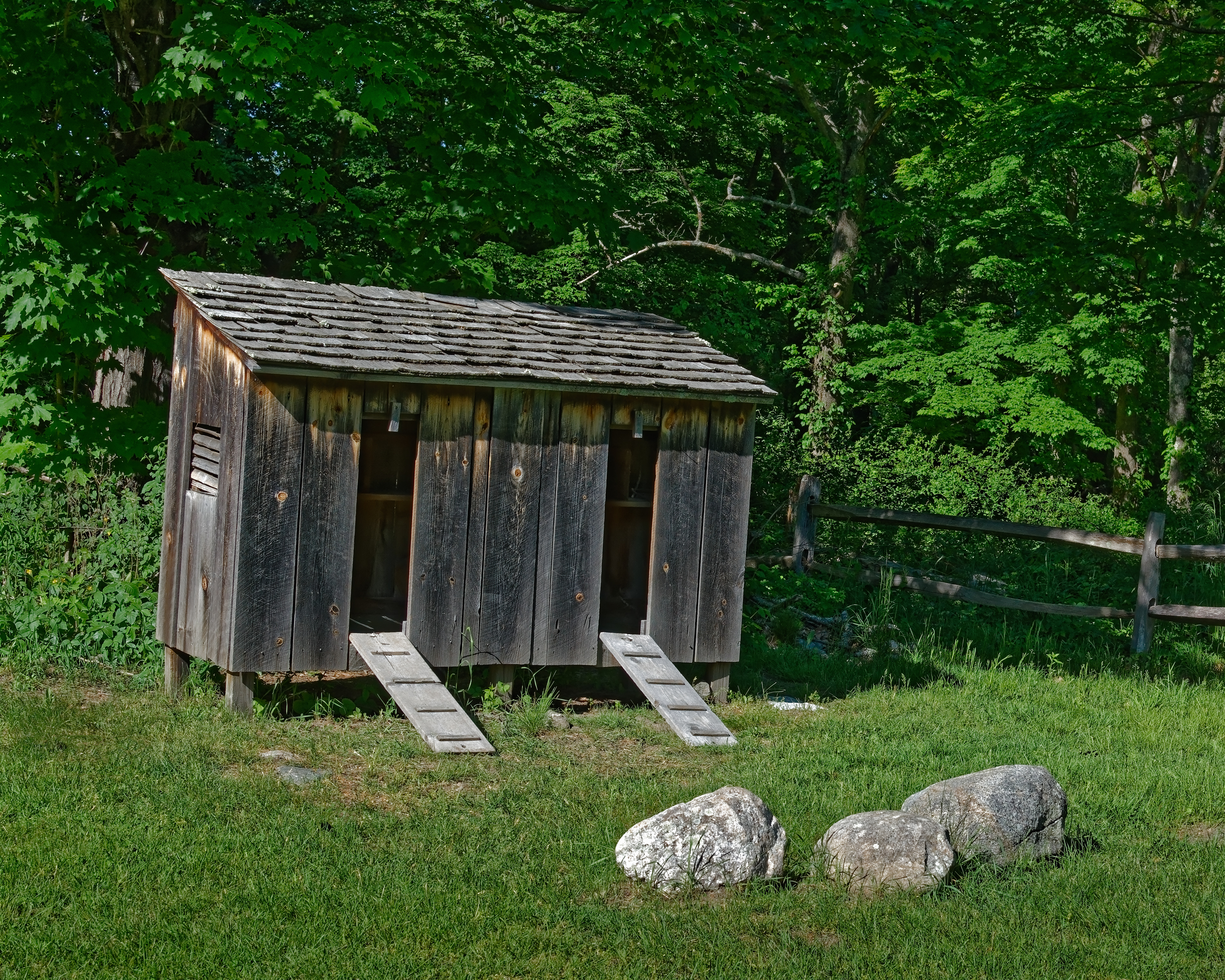 An 18th-century wood chickencoop sits empty illuminated by the setting sun on a spring evening. Short wooden runways lead up to the open doors of the shed. The roof is covered in wood shingles and the walls are made of weathered wood planks. In the background or lush green trees and a wood post fence.