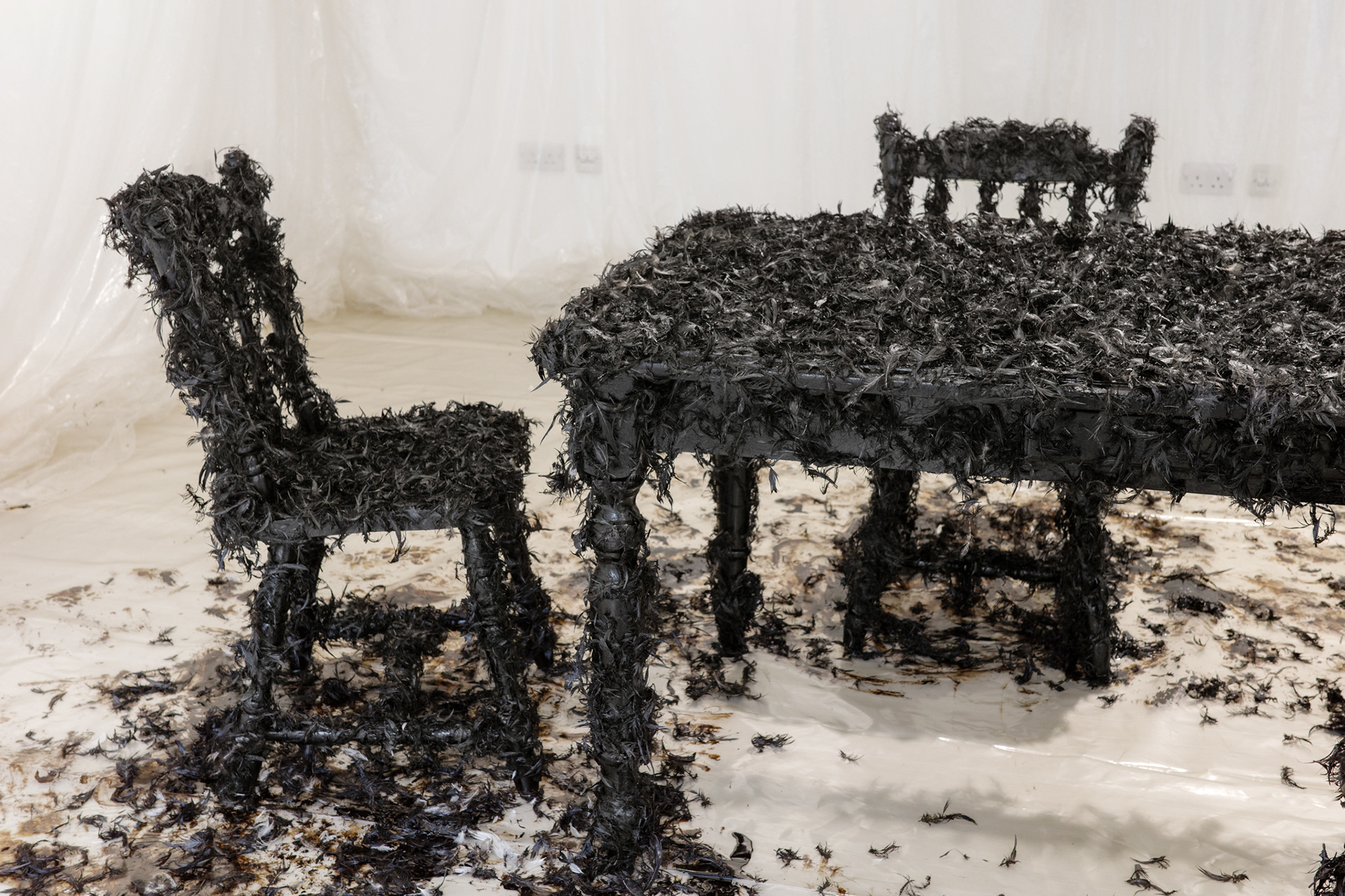 Art installation: Bitumen paint, pillow feathers, recycled wood table and chairs, plastic sheeting. Music 'Welcome to my world' written by Ray Winkler and John Hathcock and performed by Jim Reeves 1962. Installation at Brighton Centre for Contemporary Art, Brighton, UK, 2023.