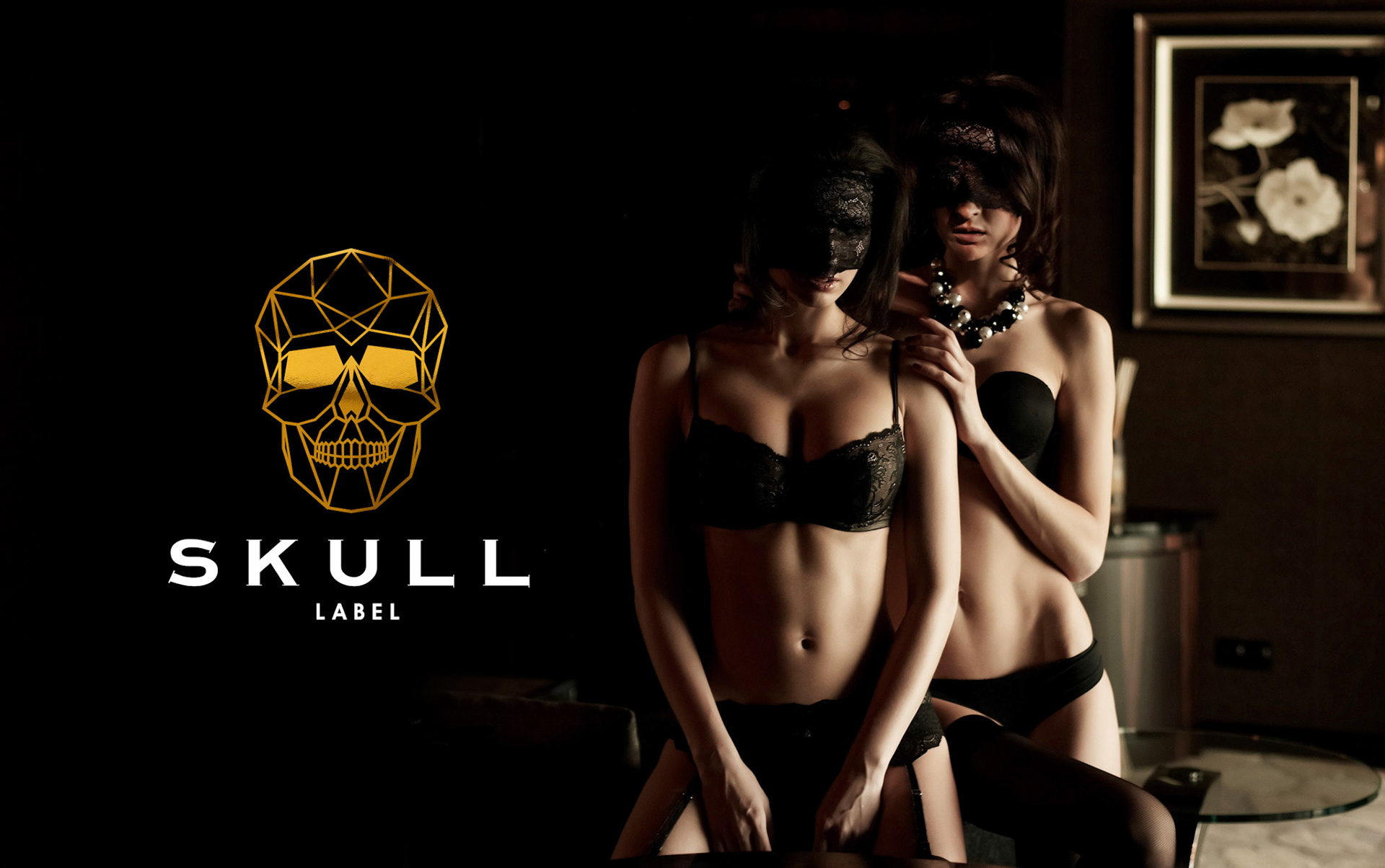 Two womans in black underwear and masks posing in shadows in luxury room.