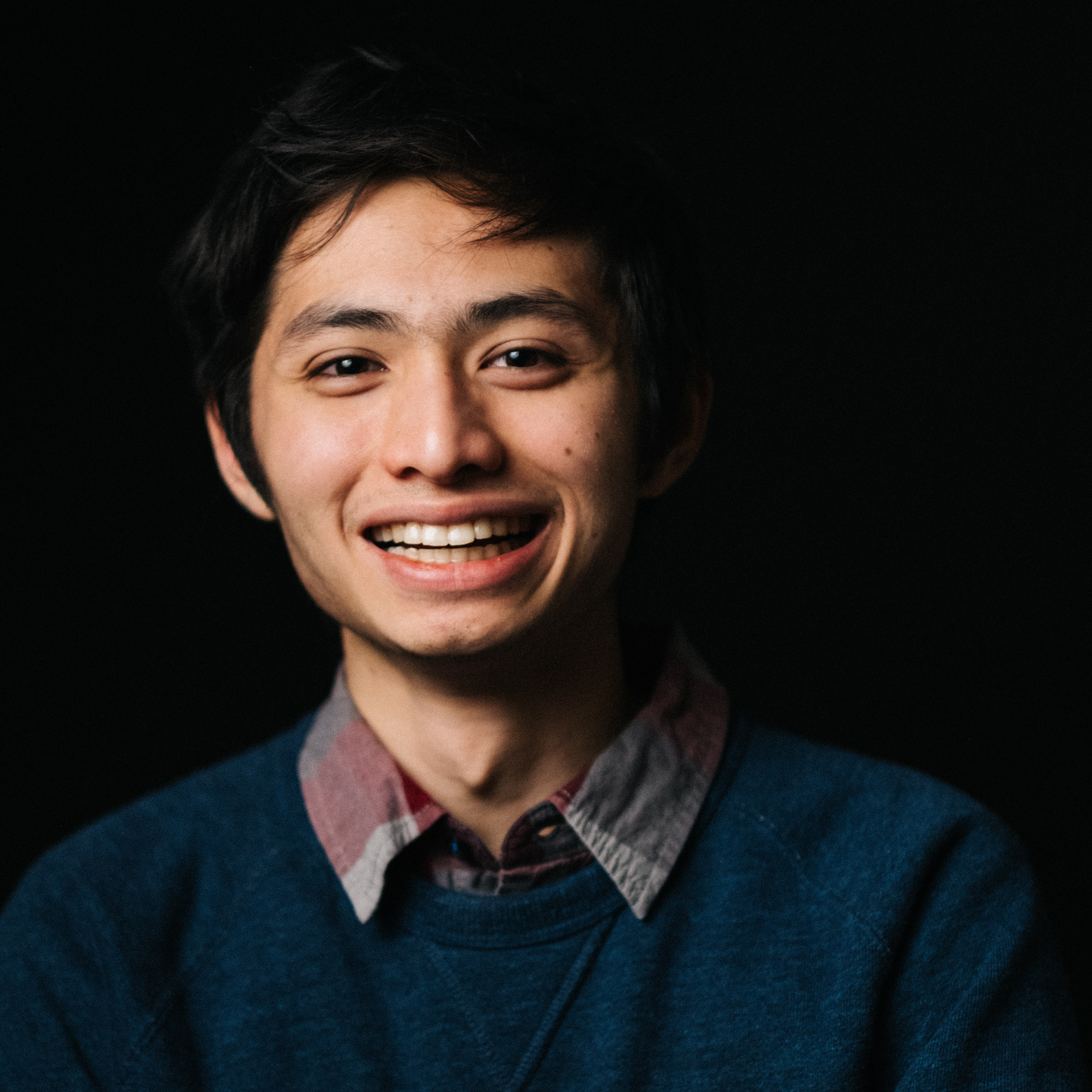 A Filipino man wearing a collared shirt and blue sweater grins for a portrait in front of a black studio background.