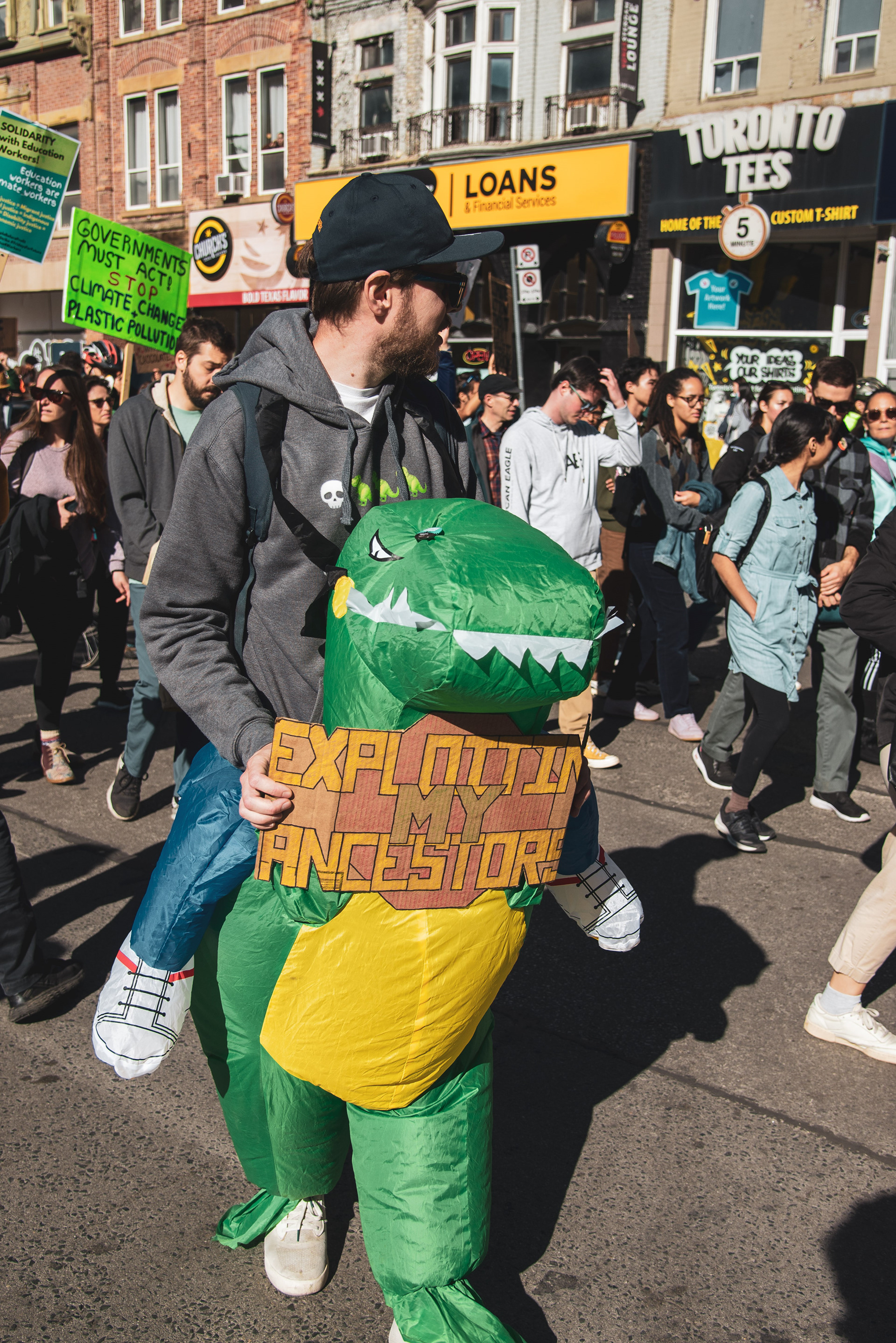 A man in a dinosaur costume holds a sign with the text, "EXPLOITING MY ANCESTORS," while marching south on Yonge Street.