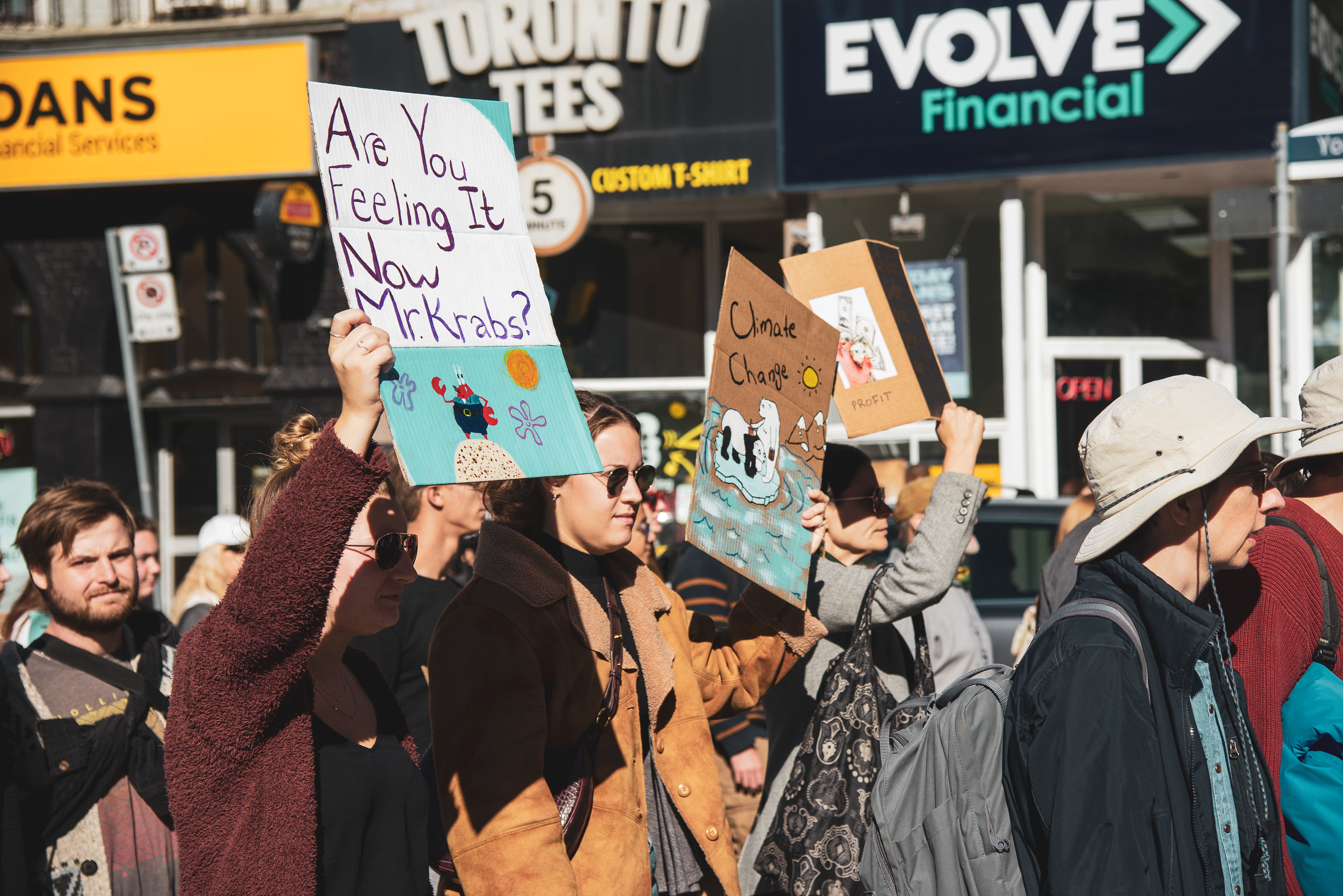 While walking down Yonge Street, a woman holds the sign with the text, "Are You Feeling It Now Mr. Krabs?" The woman next to her holds up a drawing of a polar bear and panda floating on a lone ice floe.