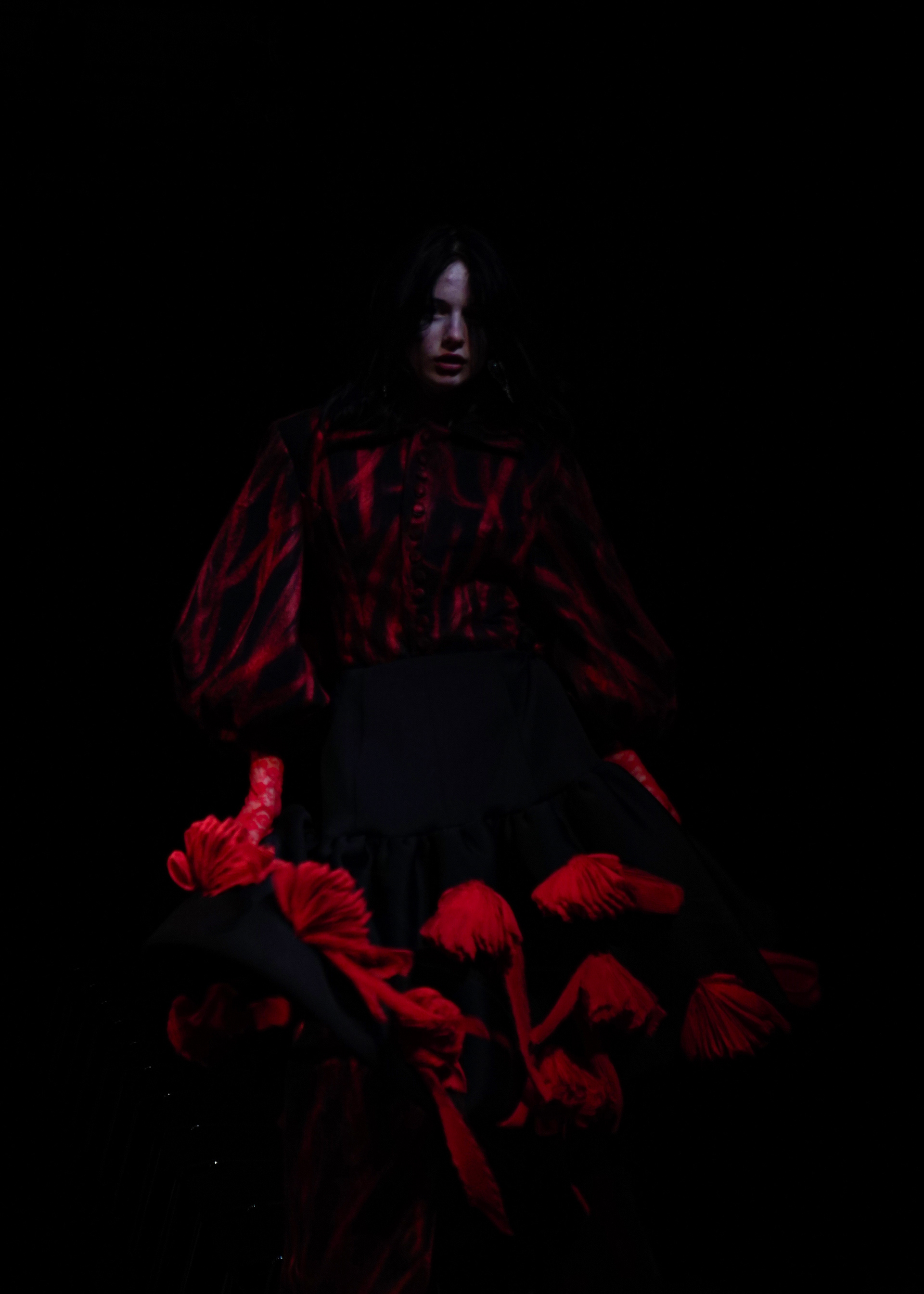 A young woman in a black and red dress stands in front of the camera, holding the frilled skirt of her dress from the ground as she walks. The image is shot incredibly dark, with much of the image blending into the shadows. The dress melts into the blackness of the background and the woman's face is seemingly cut in half, hidden behind her black hair.