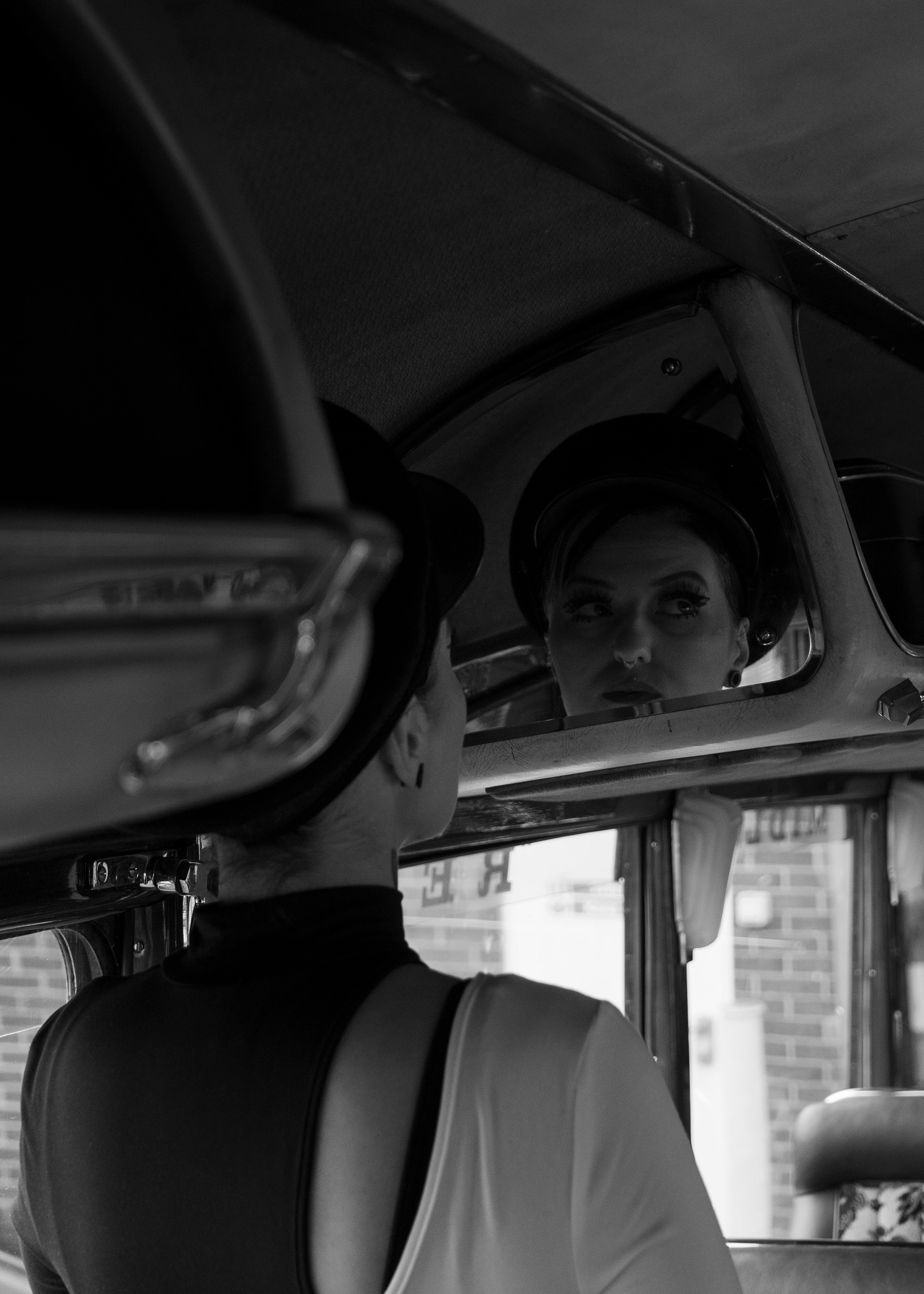 A woman stands on the interior of a vintage 1970s bus, she is stood off to the side facing a mirror built into the interior. She is dressed in a mod-inspired outfit, a black and white top paired with a large conductors cap. In her reflection we can see that she is staring off into the corner of her ceiling, a grim blank look on her face. A black and white stylisation adds to the tension in her expression.