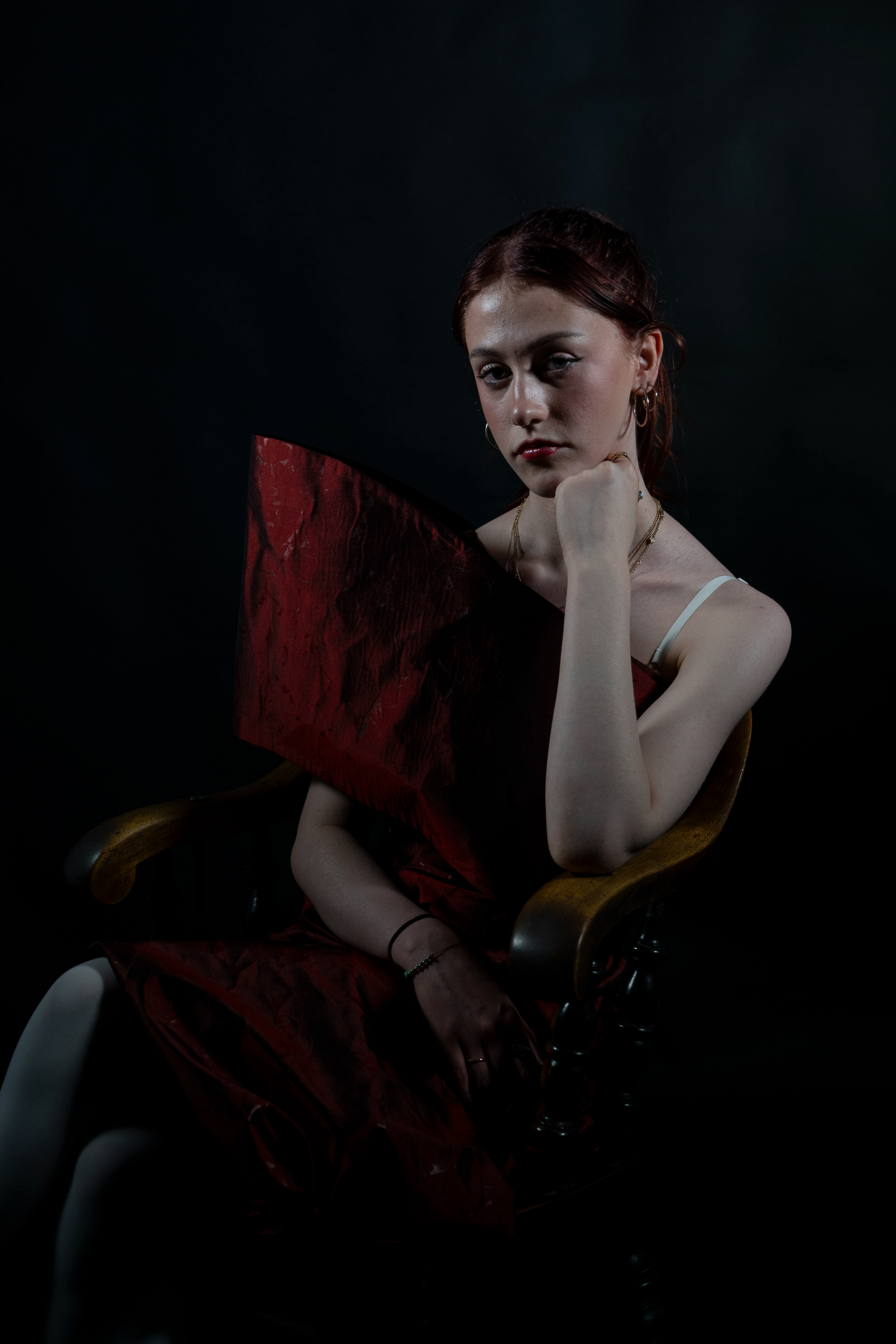 A young woman sits regally in an antique wooden chair, dramatically she rests her chin on her hand. She is dressed in a bespoke red dress by the fashion designer Drinxing Bleach, and the image is shot with minimally where only the woman's skin is brightly illuminated and the rest of the image is overtaken by shadow providing a stark contrast between her pale skin and the dark red of the garment. 