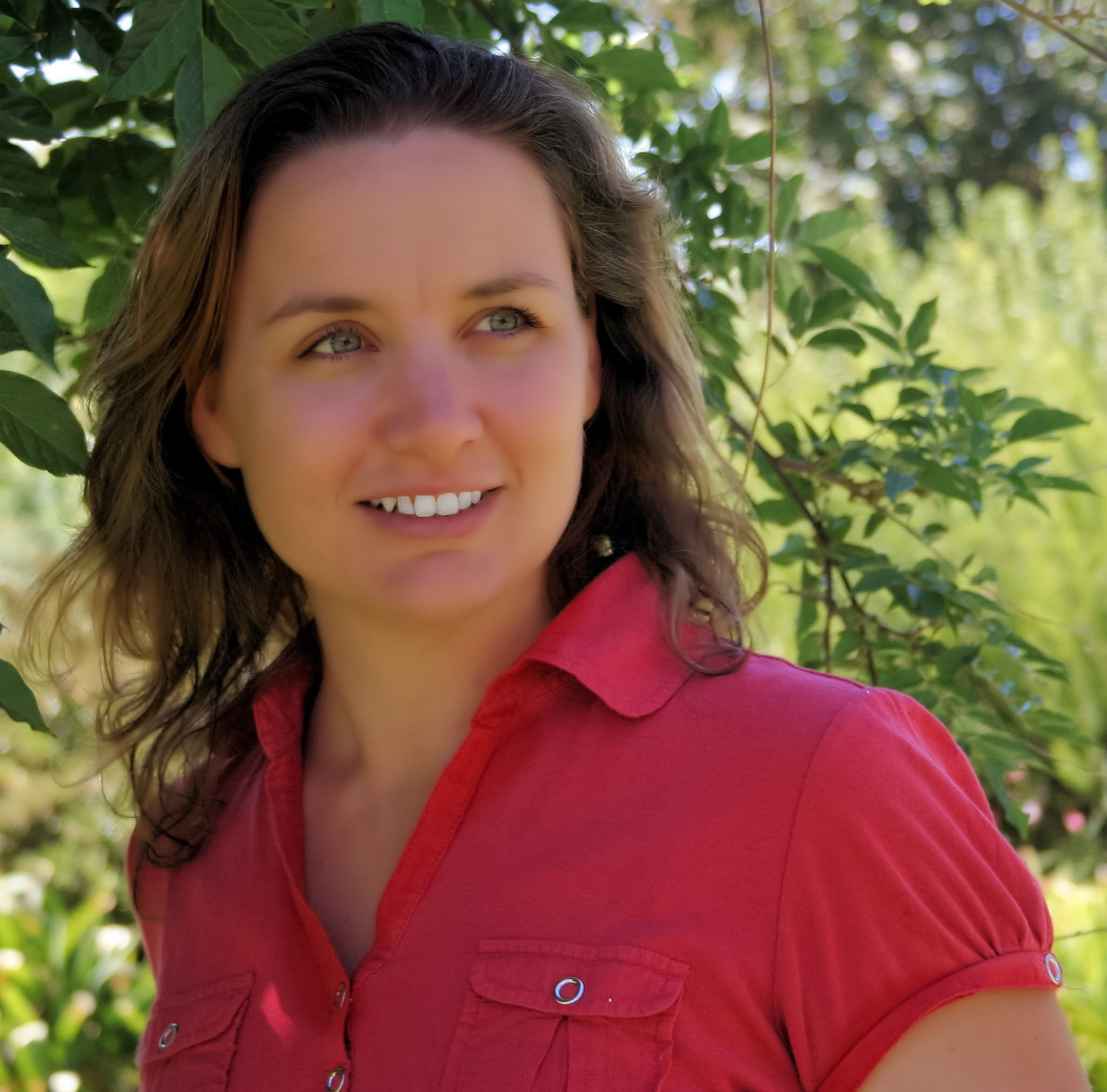 A woman with long brown hair smiles, standing outdoors amongst green foliage with natural lighting. She wears a red collared shirt.
