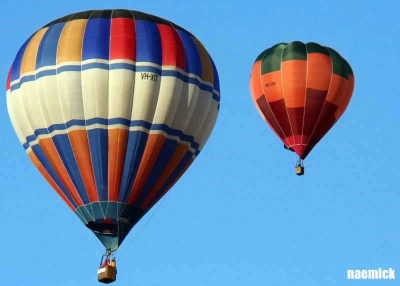 Two hot air balloons, "VH-XIT" and "VH-CSF", ascend against a clear blue sky, showcasing vibrant patterns.