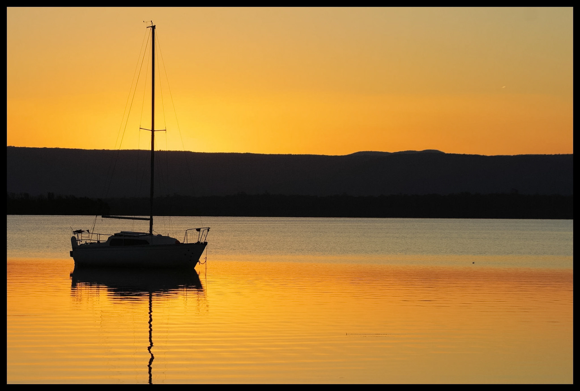 A sailboat with a mast and boom is anchored on tranquil waters, reflecting the vibrant golden sunset and silhouetted mountains.