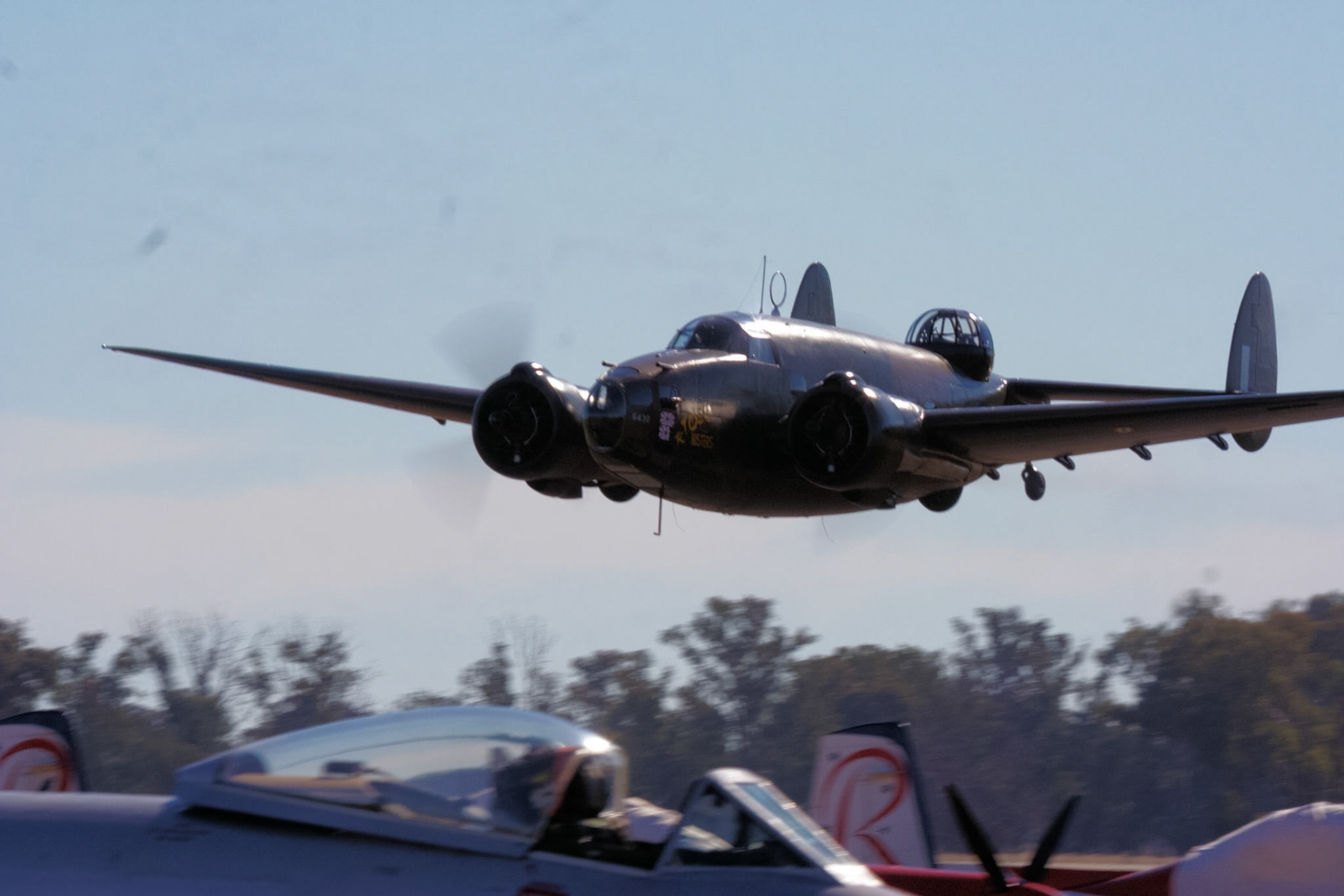 A Lockheed P2V Neptune patrol bomber, painted dark, flies low over another aircraft at an airshow event.
