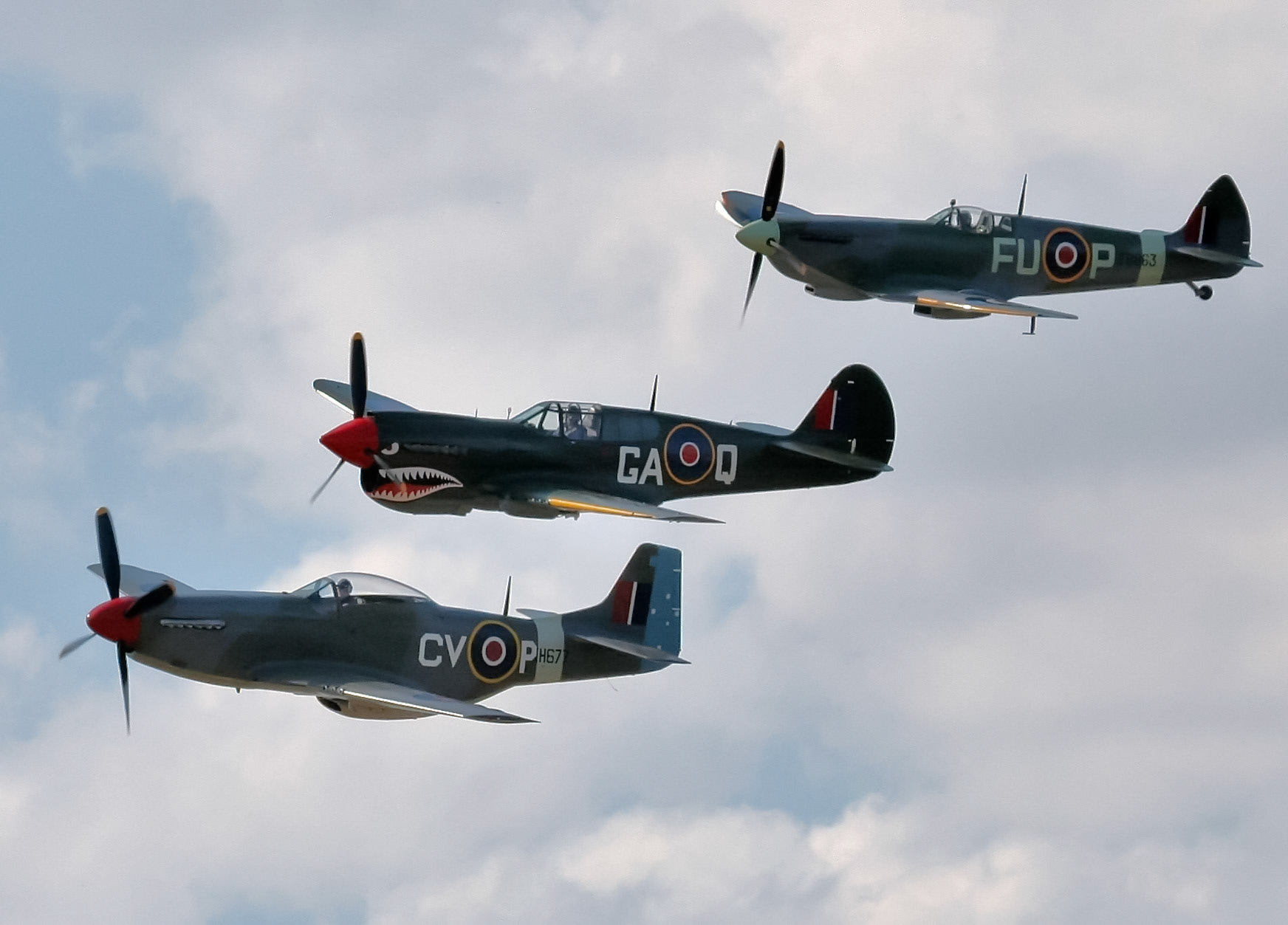 Three historic World War II fighter aircraft, a Supermarine Spitfire, Curtiss P-40 Warhawk, and North American P-51 Mustang, fly in formation.