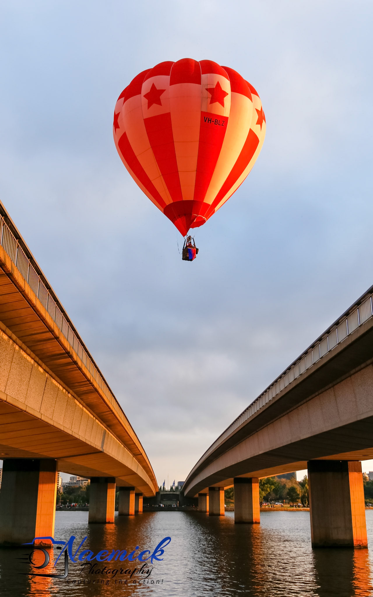 A hot air balloon with red and white stripes and stars, registered as "VH-BLZ," floats above the water in Canberra, Australia, framed by two bridges.