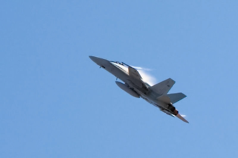 A military jet, likely a McDonnell Douglas F/A-18 Hornet, soars through a clear blue sky, leaving a contrail.