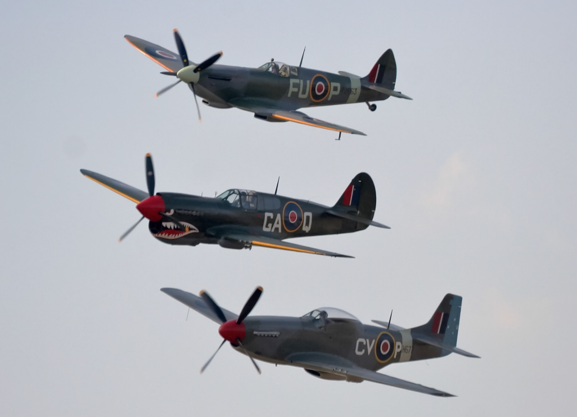 Three historic fighter aircraft, a Supermarine Spitfire, Curtiss P-40 Warhawk, and North American P-51 Mustang, fly in formation.