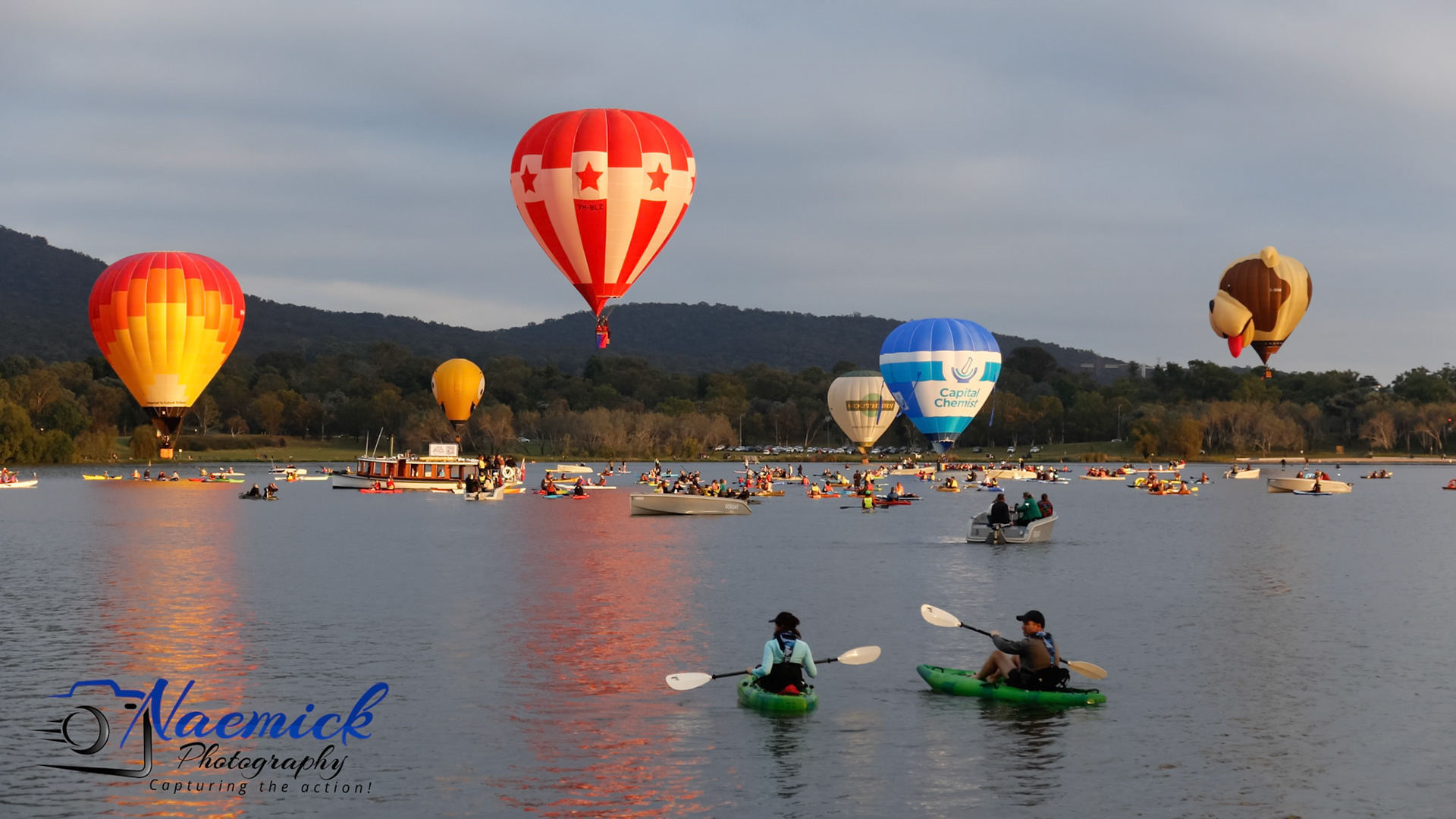 Hot air balloons float over a lake in Canberra, Australia, with many boats and kayaks on the water. "Naemick Photography Capturing the action!"