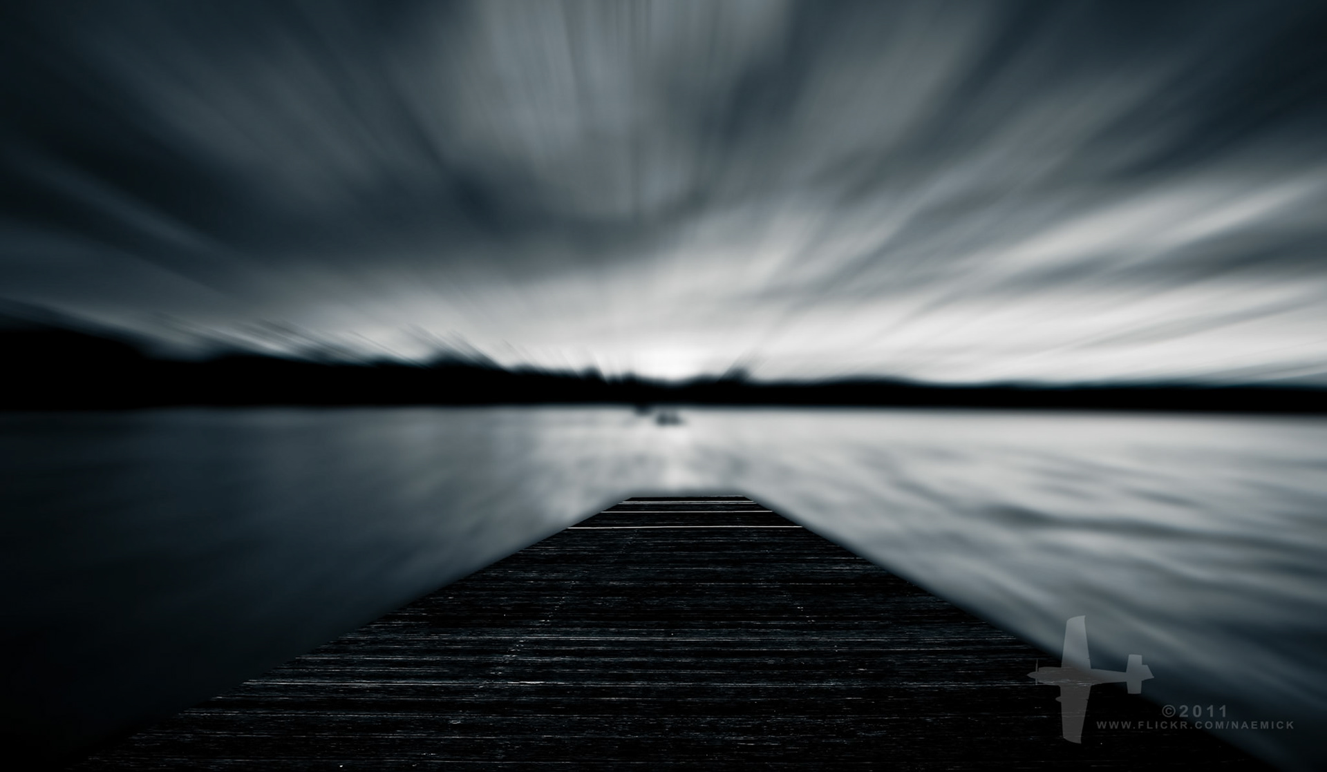 A long exposure photograph shows a wooden pier extending into calm water under a dramatically streaked sky. Distant land is visible across the water. The image features a watermark "©2011 WWW.FLICKR.COM/NAEMICK".