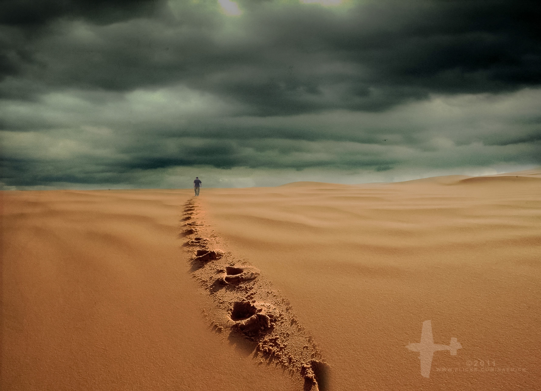 A lone figure walks across a vast desert landscape, leaving footprints in the sand under a dramatic, cloudy sky.