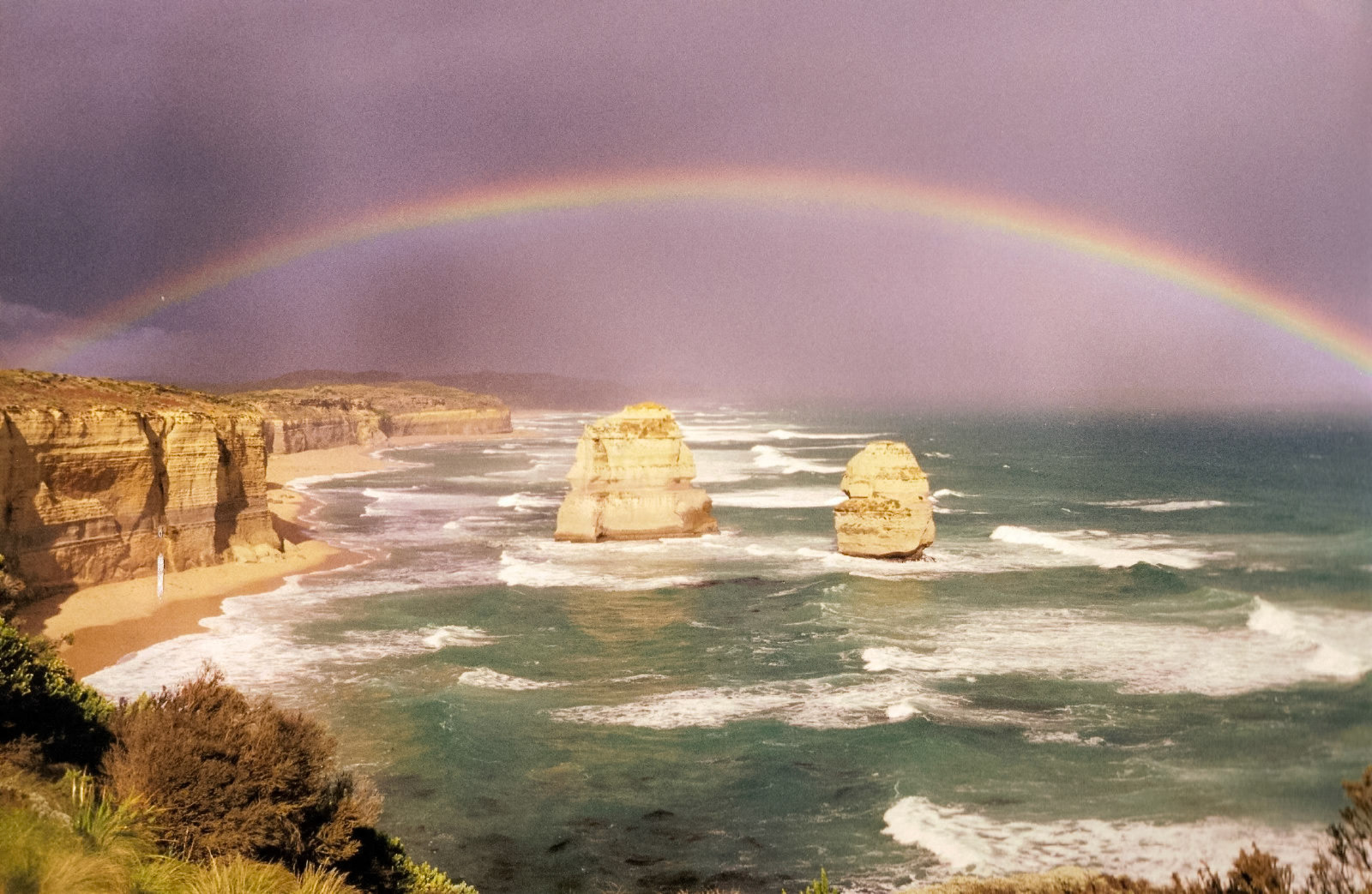 A scenic view of the Twelve Apostles rock formations along the Victorian coastline, with waves crashing on the shore and a rainbow spanning the sky.