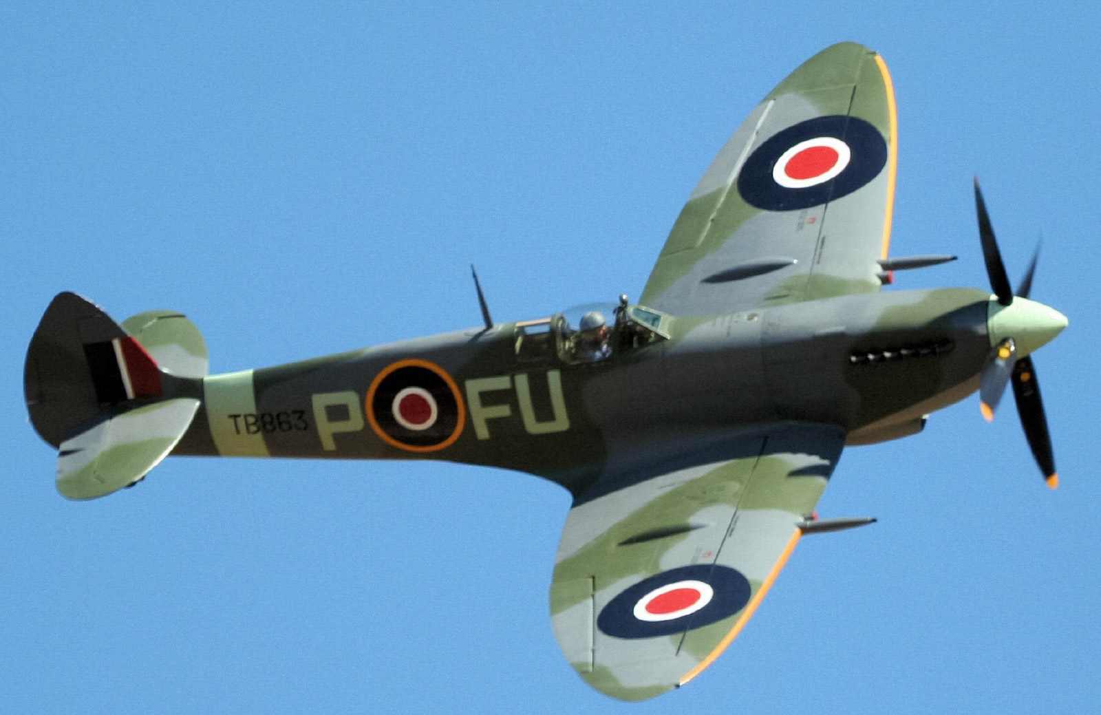A vintage Supermarine Spitfire aircraft, with registration “TB863” and codes “POFU”, flies against a clear blue sky. The plane displays Allied roundels on its wings and fuselage.