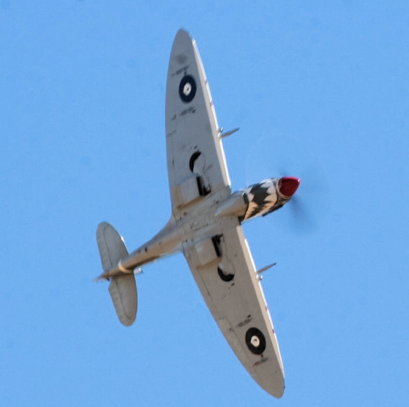 A vintage Supermarine Spitfire aircraft, with a red propeller nose cone and black and white striped engine cowling, in flight against a clear blue sky. "G-IXD" is visible on the underside of the wing. It is an iconic World War II British fighter.
