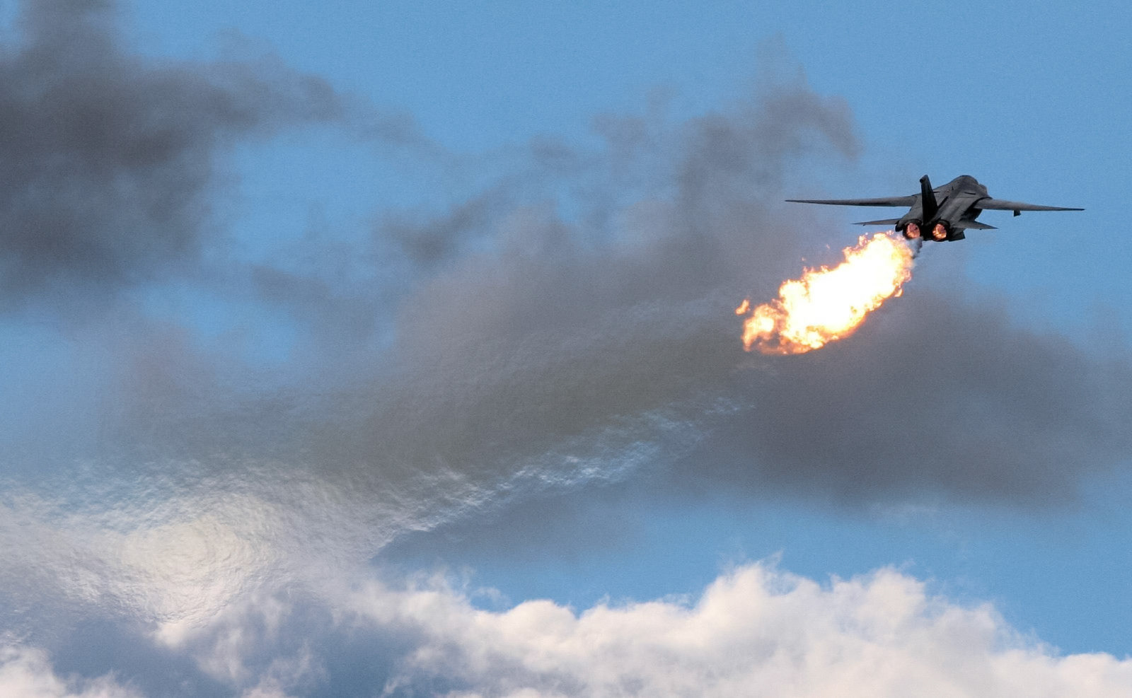 A military jet with afterburners engaged shoots a large plume of fire into the sky against a cloudy background.