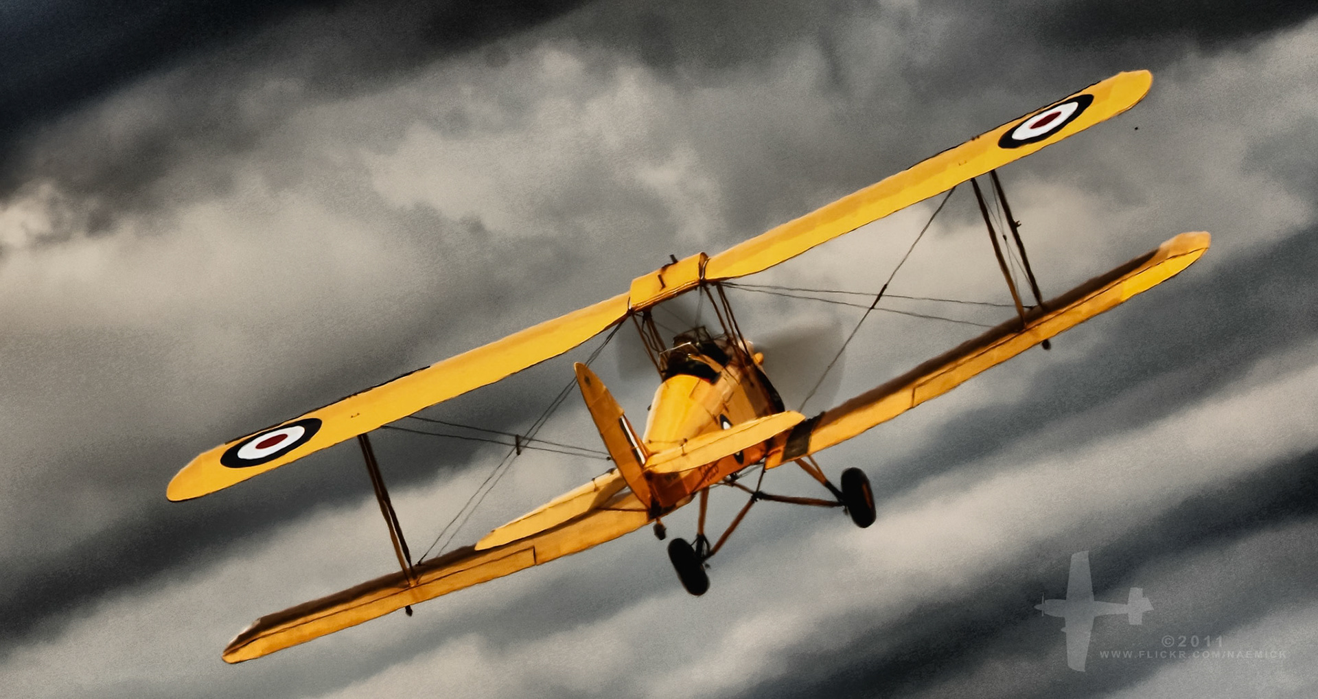 A vintage yellow biplane, identified as a de Havilland Tiger Moth, performs an aerial maneuver against a cloudy sky. "©2011 WWW.FLICKR.COM/NAZIMIOK"
