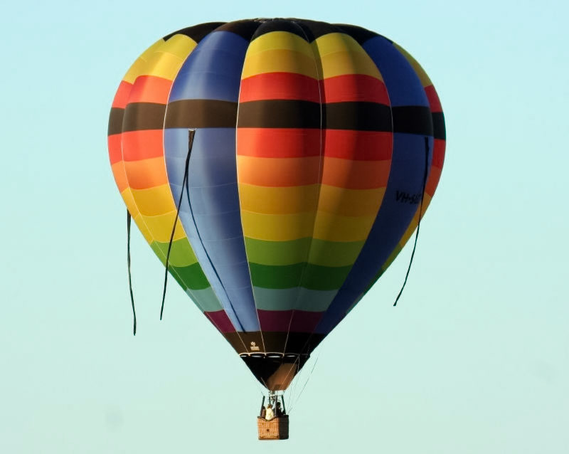 A vibrantly colored hot air balloon, featuring prominent stripes in red, orange, yellow, green, blue, and black, ascends into a clear, light blue sky. The registration number "VH-SHA" is visible.