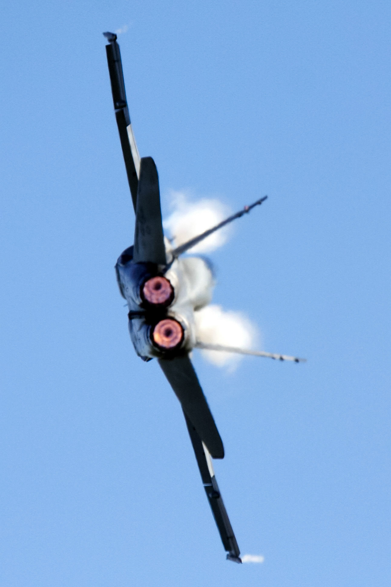 A military jet with glowing afterburners and exhaust plumes against a clear blue sky, seen from below.
