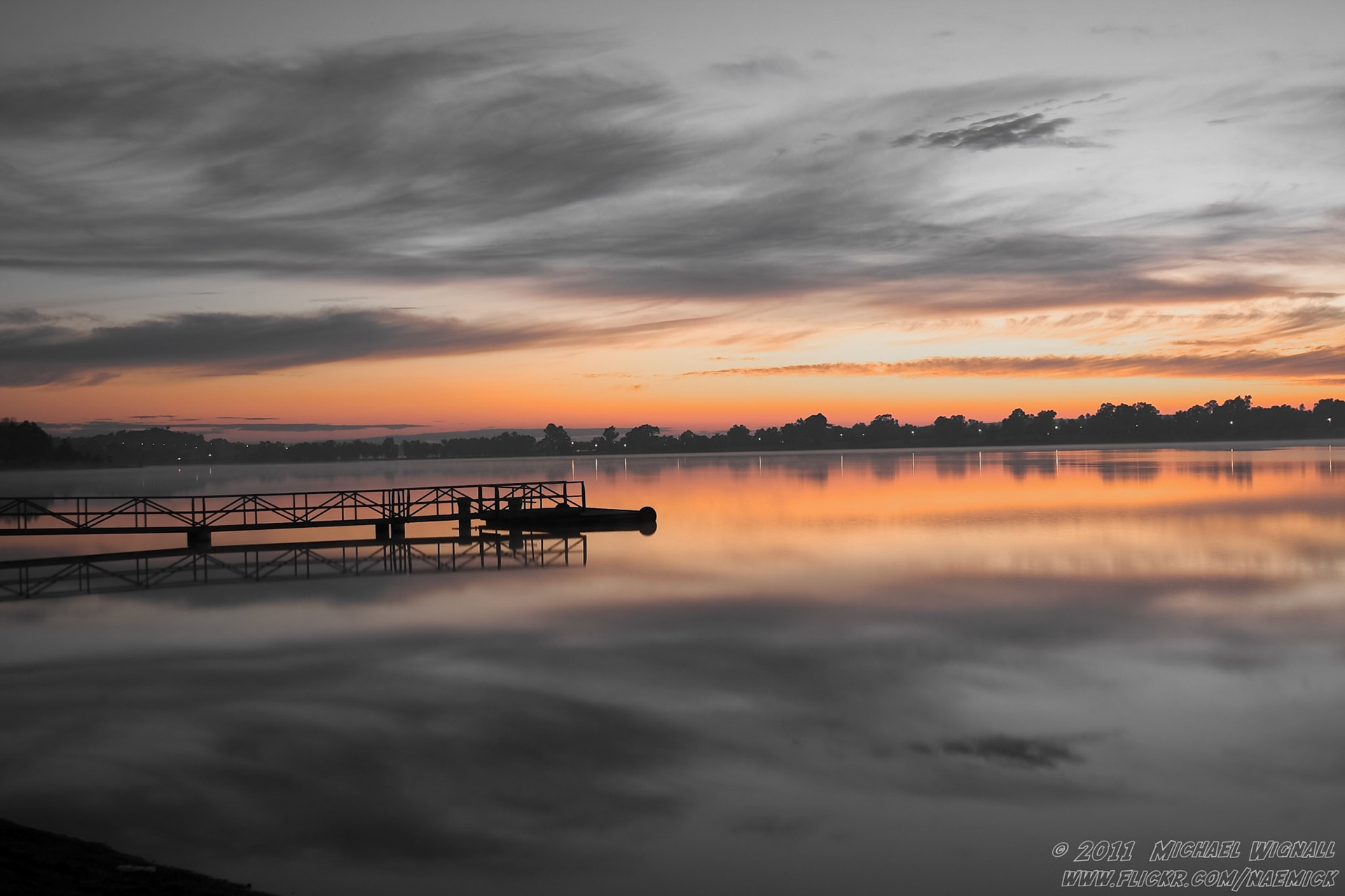 A tranquil dawn scene with a pier extending into still waters, reflecting the vibrant orange sky above.