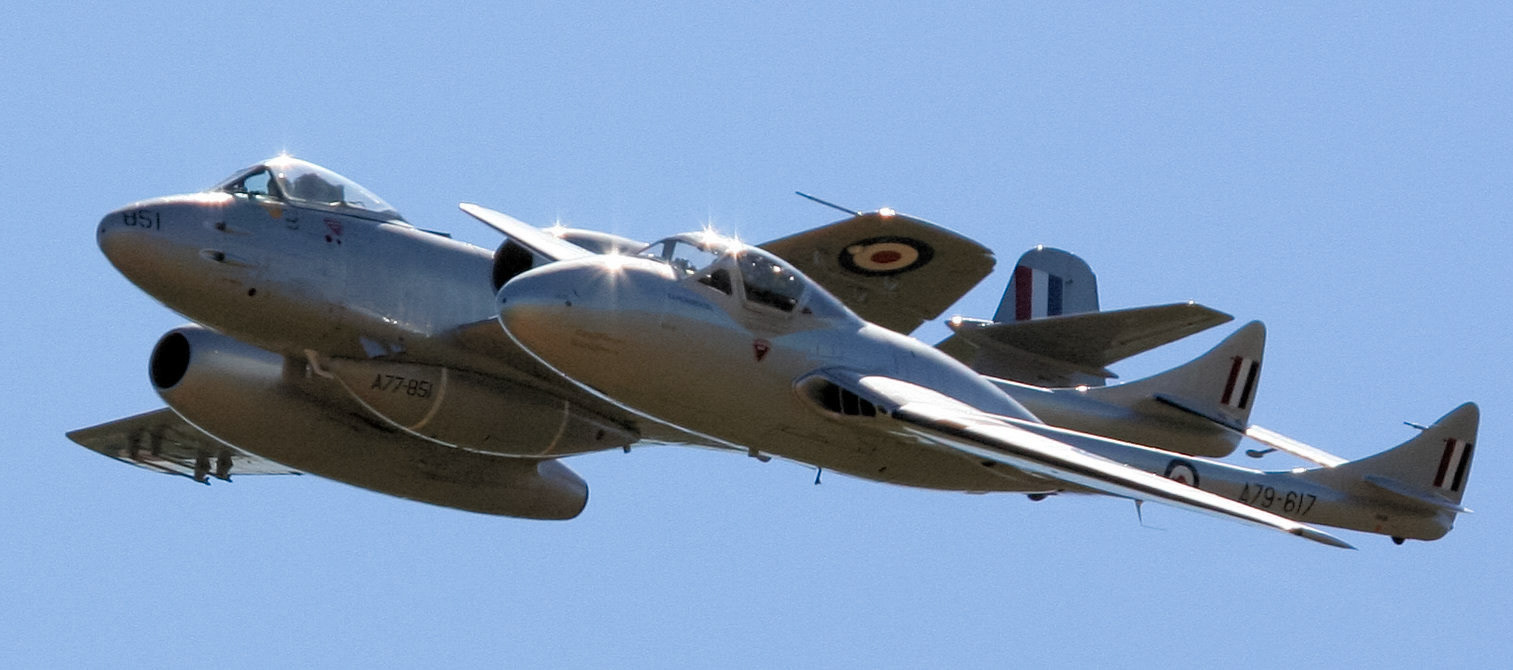 Two de Havilland Vampire jet aircraft, registrations "A77-851" and "A79-617", fly in formation against a clear blue sky. "851" is visible on the nose.
