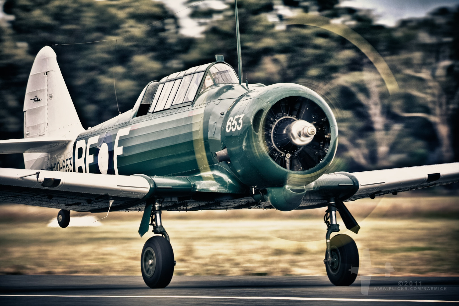 A vintage North American T-6 Texan aircraft, painted in military green, is captured with its propeller spinning on a runway. The plane displays the numbers "653" and letters "BFOF".