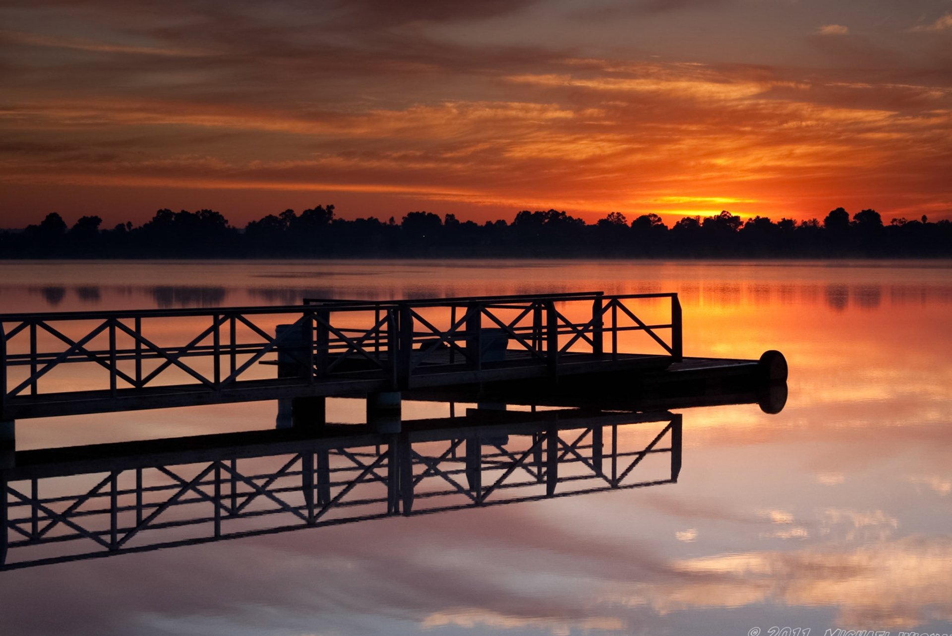A tranquil lake scene with a wooden dock stretching into the calm water, reflecting the vibrant orange sunset sky.