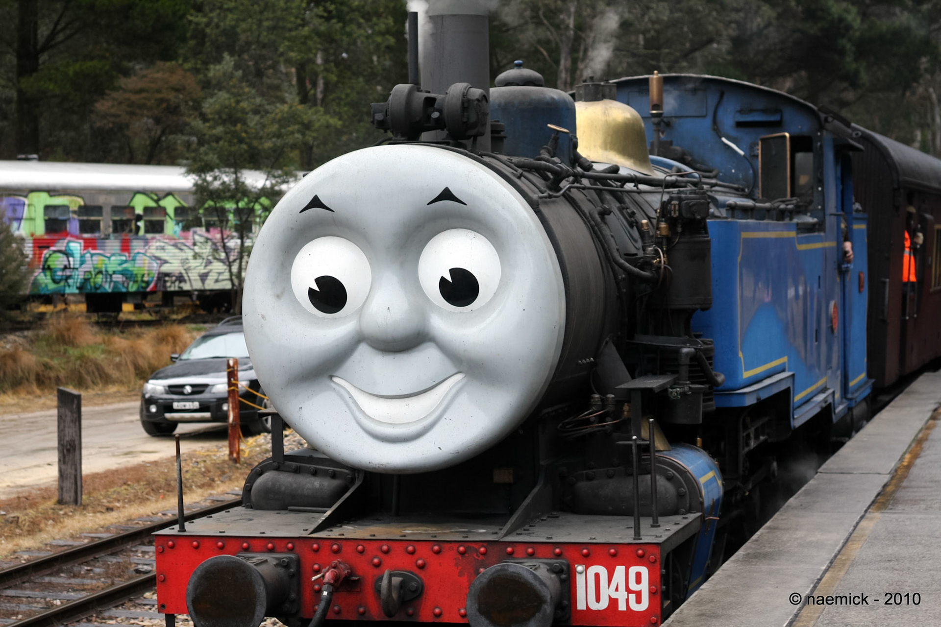 A blue and black steam locomotive, decorated with a "Thomas the Tank Engine" face, sits on tracks next to a platform, displaying the number "1049".