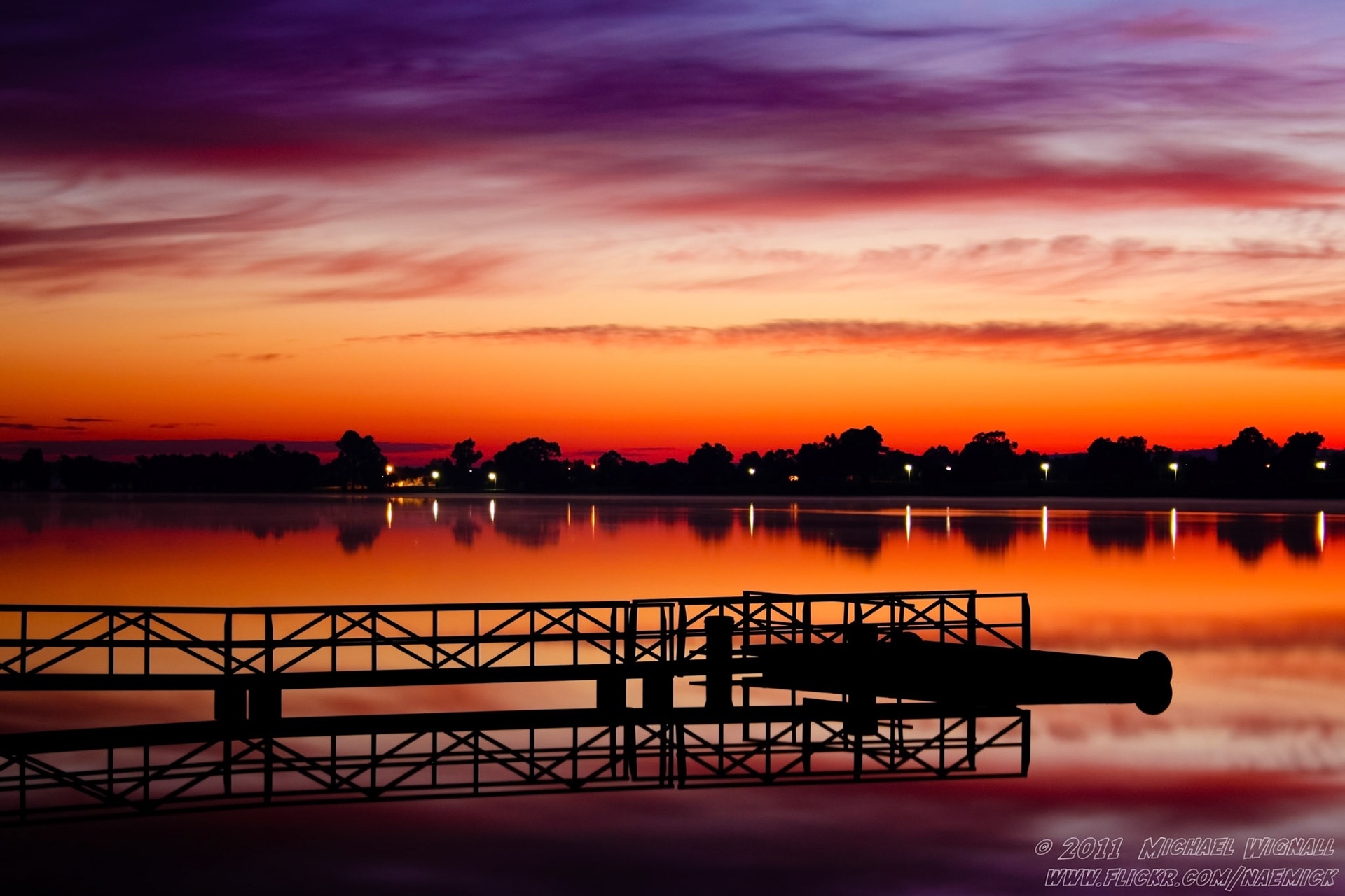 A tranquil lake reflects the vibrant orange, red, and purple hues of a sunset sky. A dark pier and boat are silhouetted.