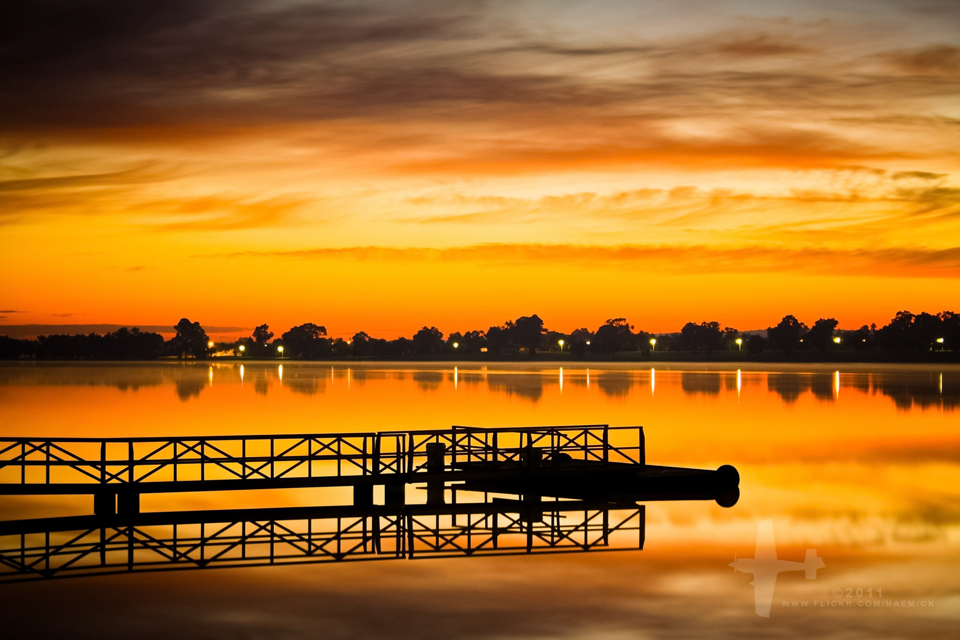 A tranquil water scene features a silhouetted pier reflecting on the calm surface under an intensely orange and yellow sky.