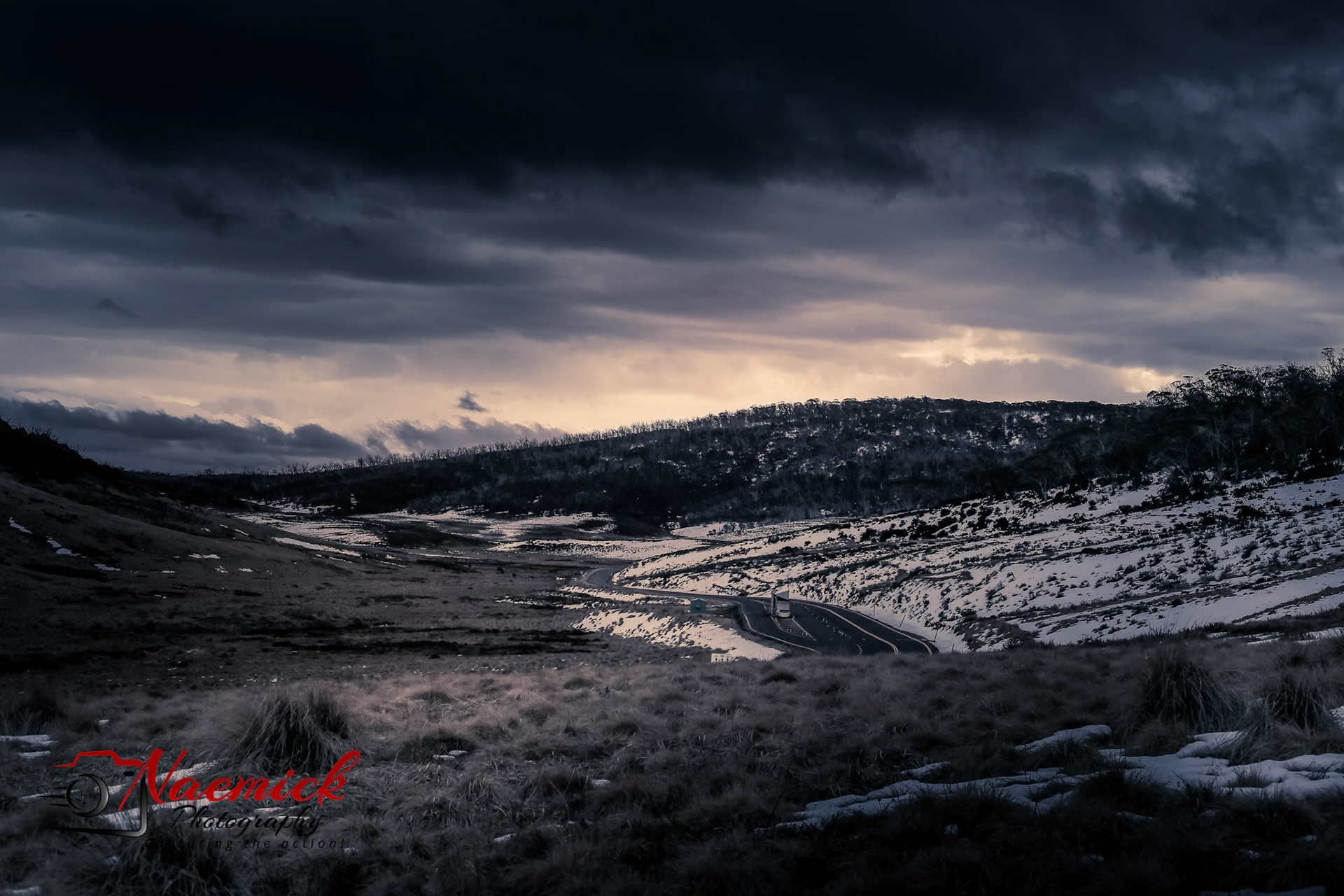 A winding road traverses a snow-dusted landscape under dramatic, cloudy skies in New South Wales, Australia.
