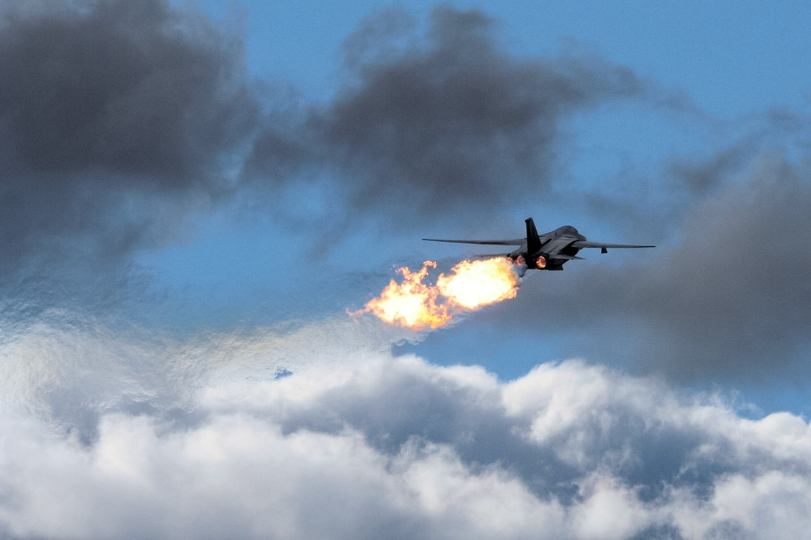 A fighter jet with afterburners engaged ascends rapidly into a partly cloudy sky, leaving a trail of fire.