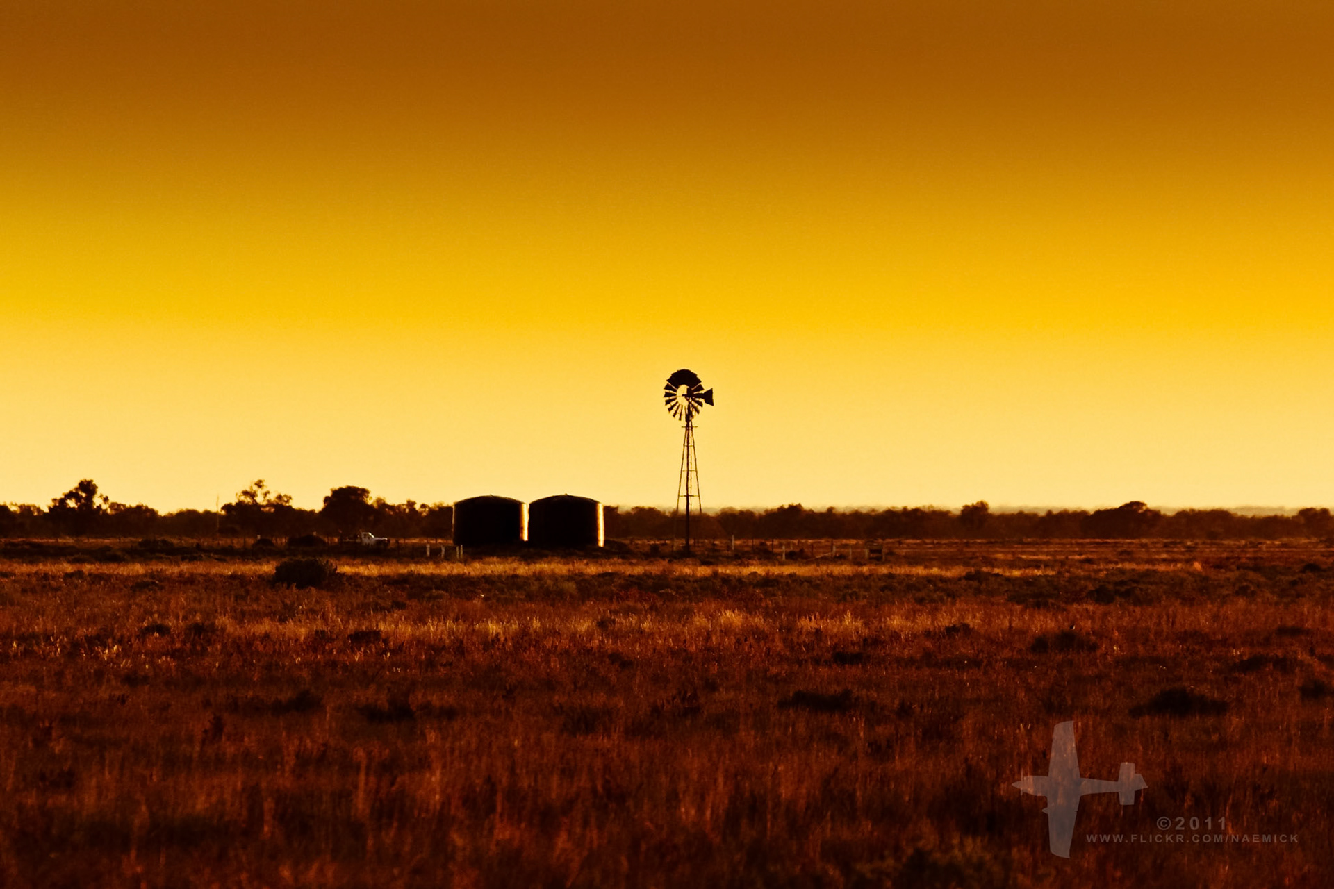 A silhouetted windmill and two water tanks stand against a vibrant orange and yellow sky at sunset, in a vast, dry field.