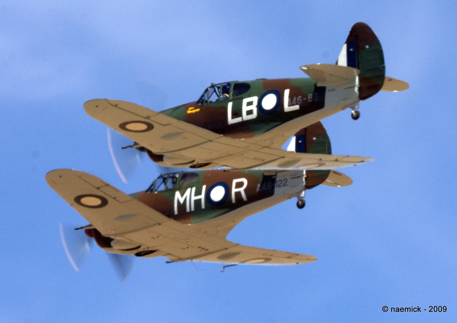 Two World War II era P-40 Kittyhawk aircraft, “A46-63” and “A46-122”, perform an aerial display against a clear blue sky.