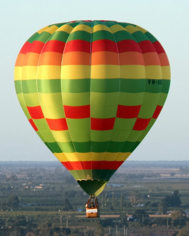 A colorful hot air balloon with red, yellow, orange, and green horizontal stripes, displaying the registration "VH-SSK", ascends above a green landscape.