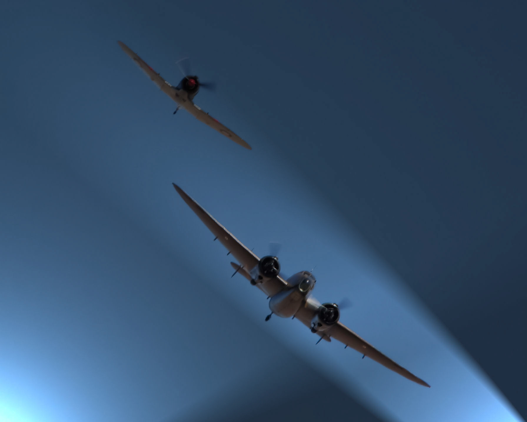 Two vintage propeller airplanes are captured in flight against a clear blue sky, with a spotlight beam illuminating one.