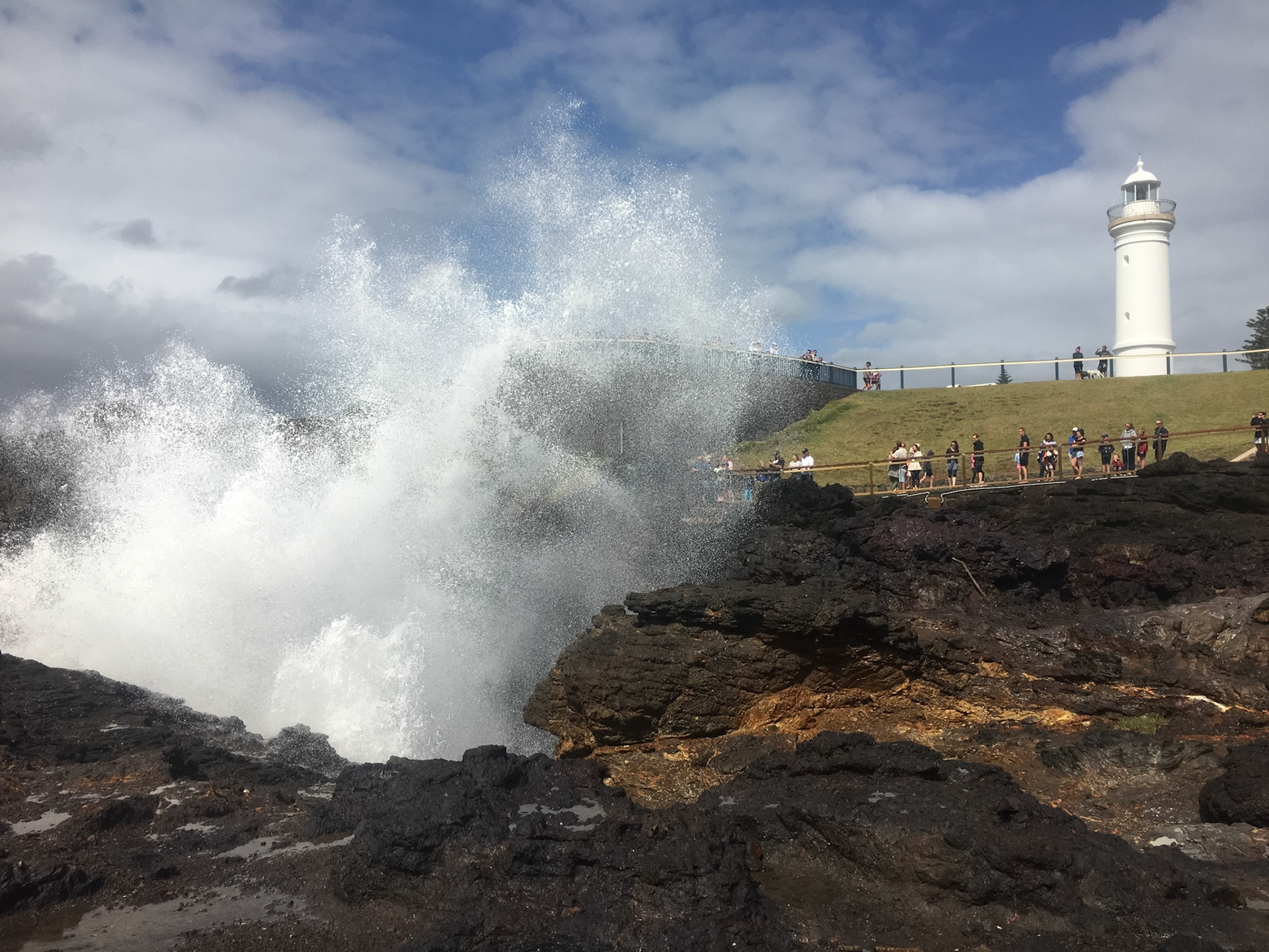 A crowd of people observe the Kiama Blowhole erupting, with the historic Kiama Lighthouse standing on a hill in the background.