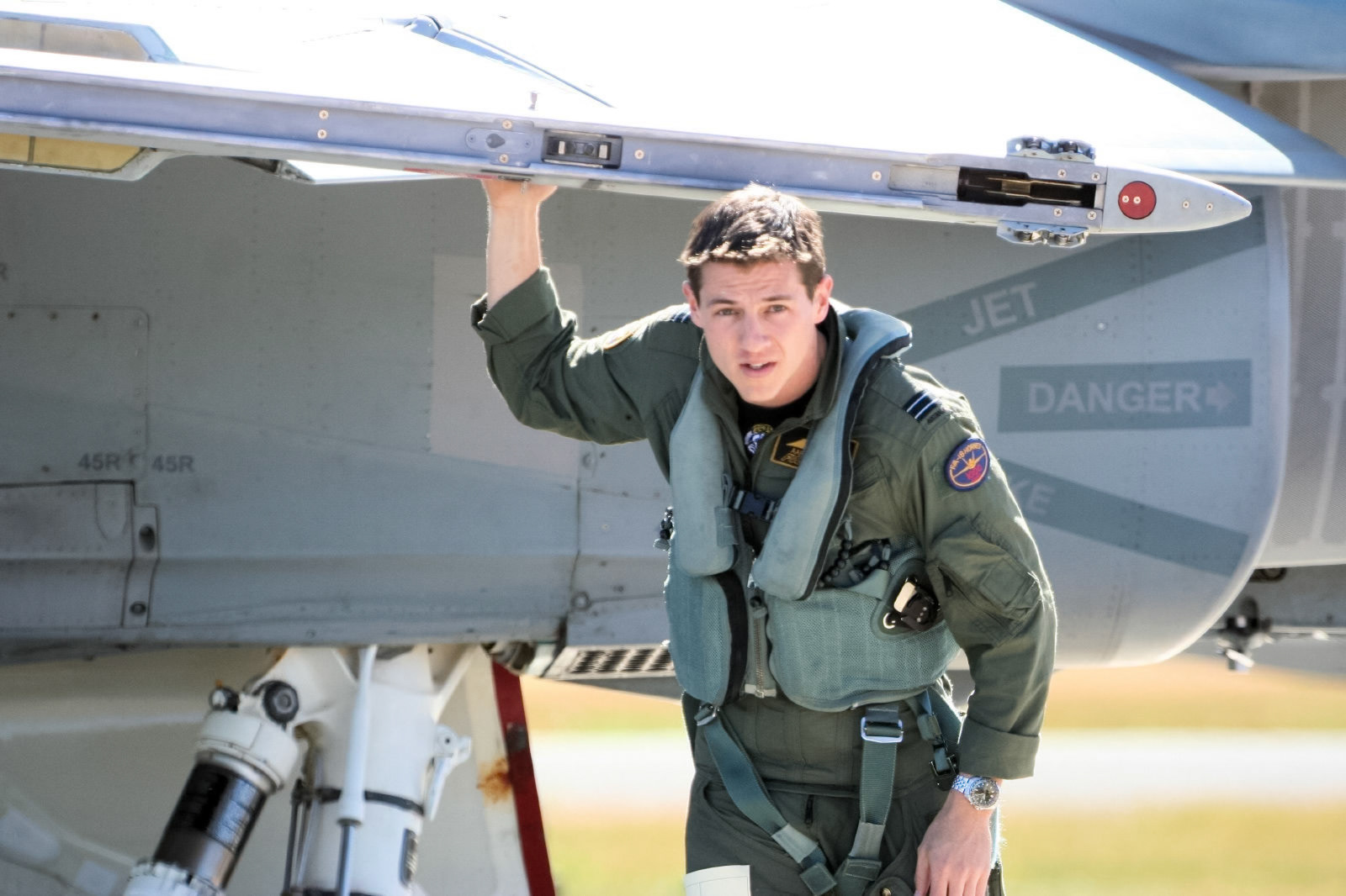 A pilot in a green flight suit and life vest stands beside a military jet with "JET DANGER" visible.