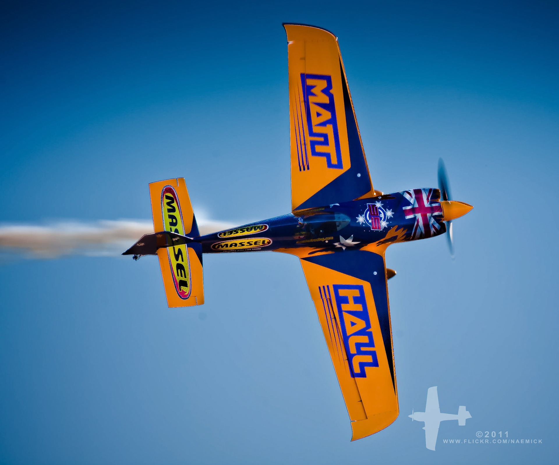 An aerobatic airplane, with "MATT HALL" and "MASSEL" emblazoned, performs a maneuver, leaving a smoke trail in the sky.