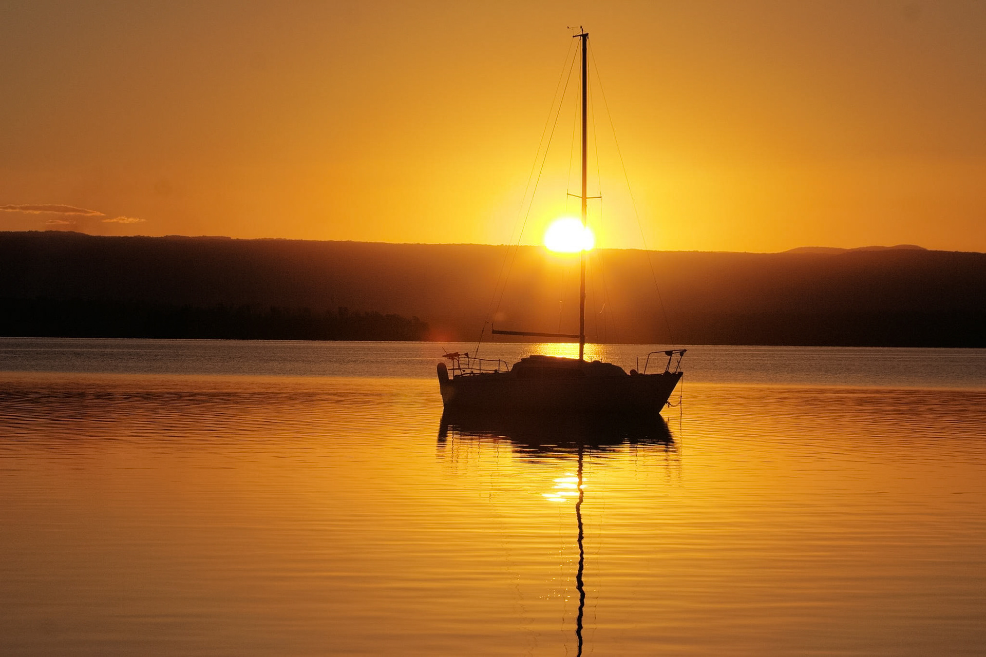 A tranquil sunset over water, silhouetting a sailboat with its mast directly in front of the sun.