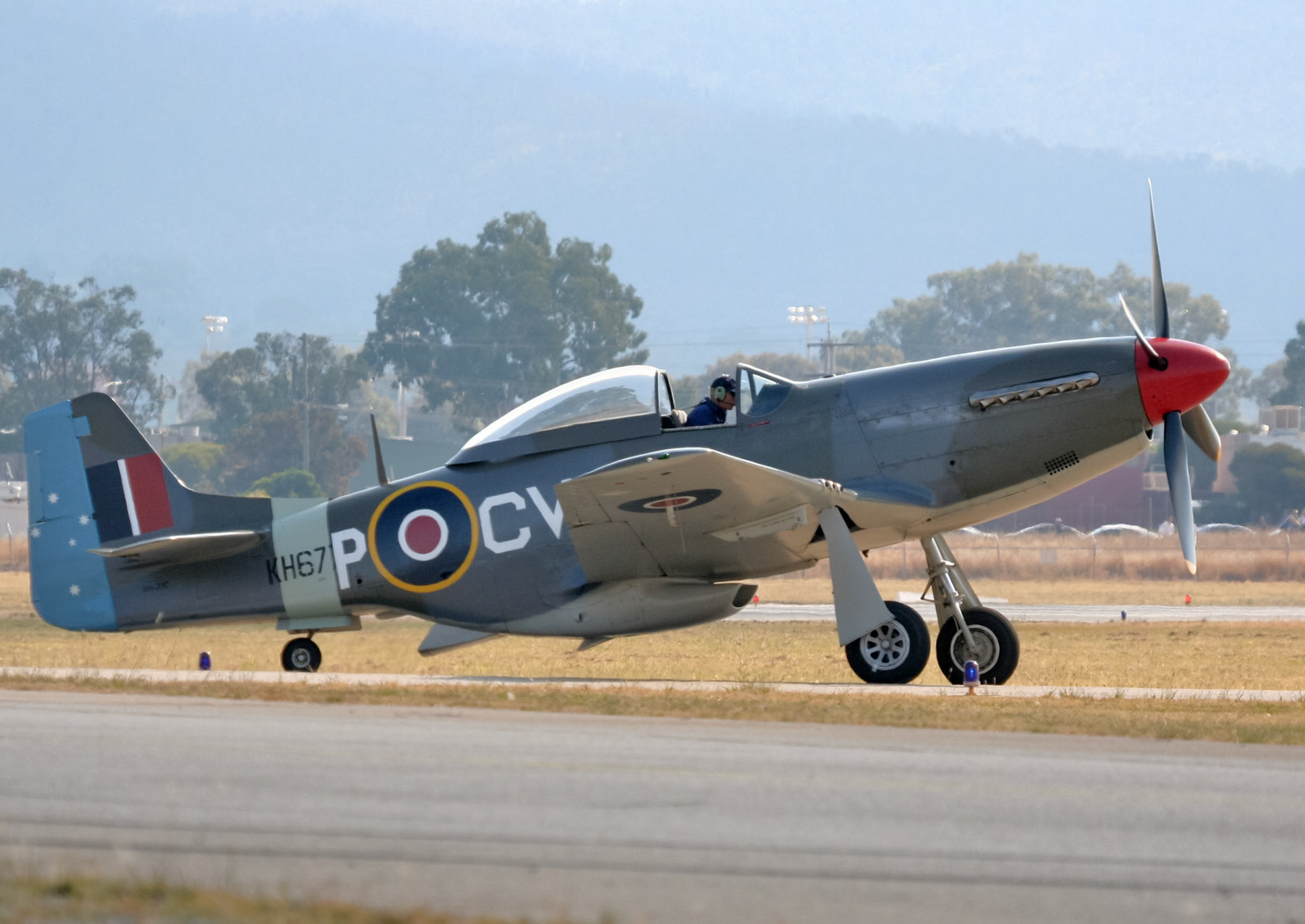 A vintage North American P-51 Mustang aircraft, with tail number "KH67 POCV", is positioned on a tarmac, preparing for flight.