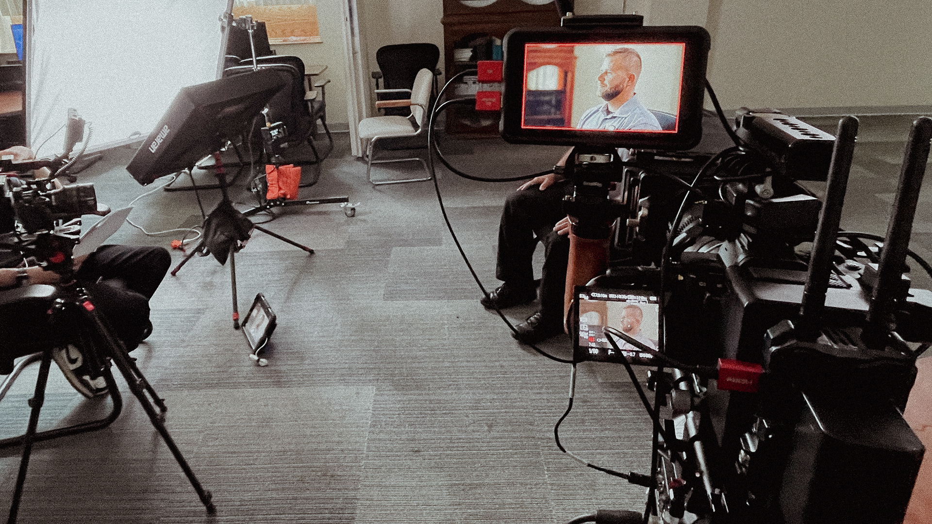Behind-the-scenes of a video shoot with multiple cameras, lighting equipment, cables, and monitors displaying a man being interviewed.